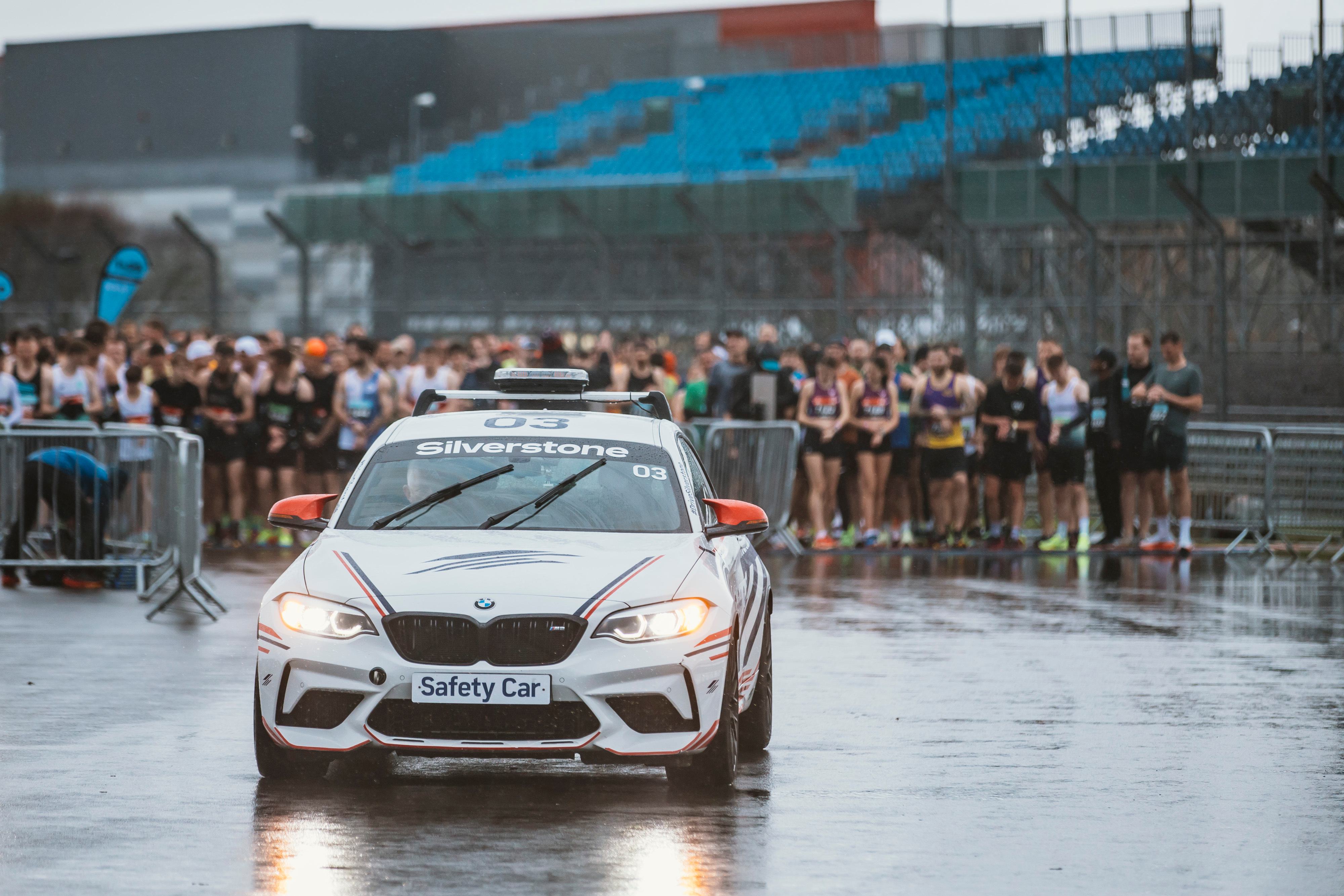 A BMW safety car with "Silverstone" written on it leads a group of runners at an outdoor race event. The ground is wet, and the scene includes a stadium in the background.