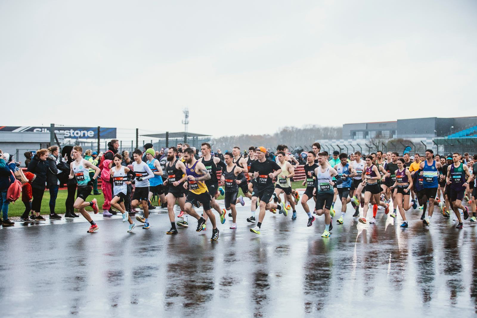 A large group of runners starts a race on a wet track, with a crowd of spectators in the background. The runners are wearing athletic gear, and the sky is overcast.