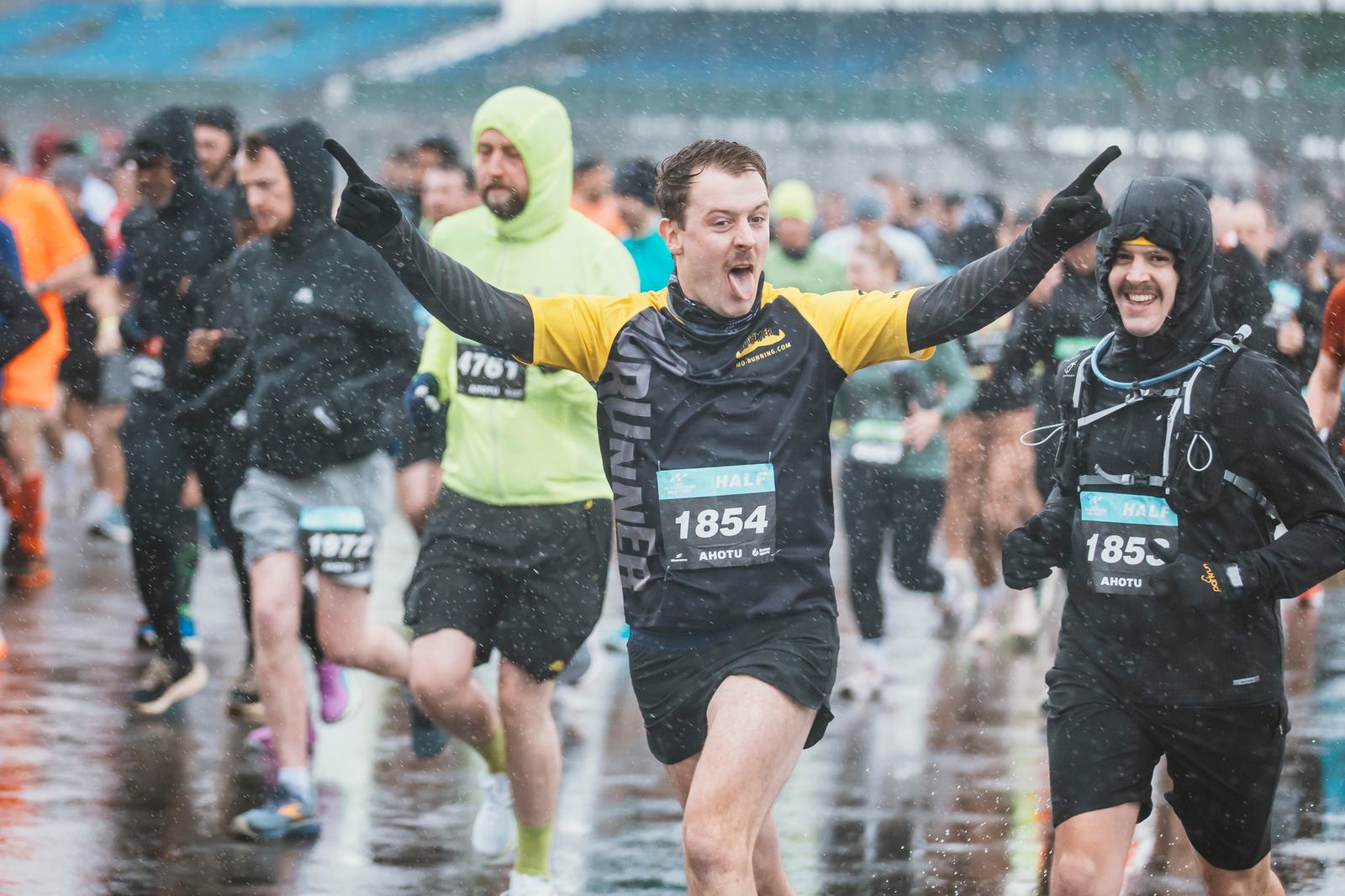 Runners participating in a race on a rainy day. A man in a black and yellow outfit is at the center, smiling with his arms raised in celebration. Other runners are behind him, some wearing rain gear. The ground is wet, reflecting the overcast sky.