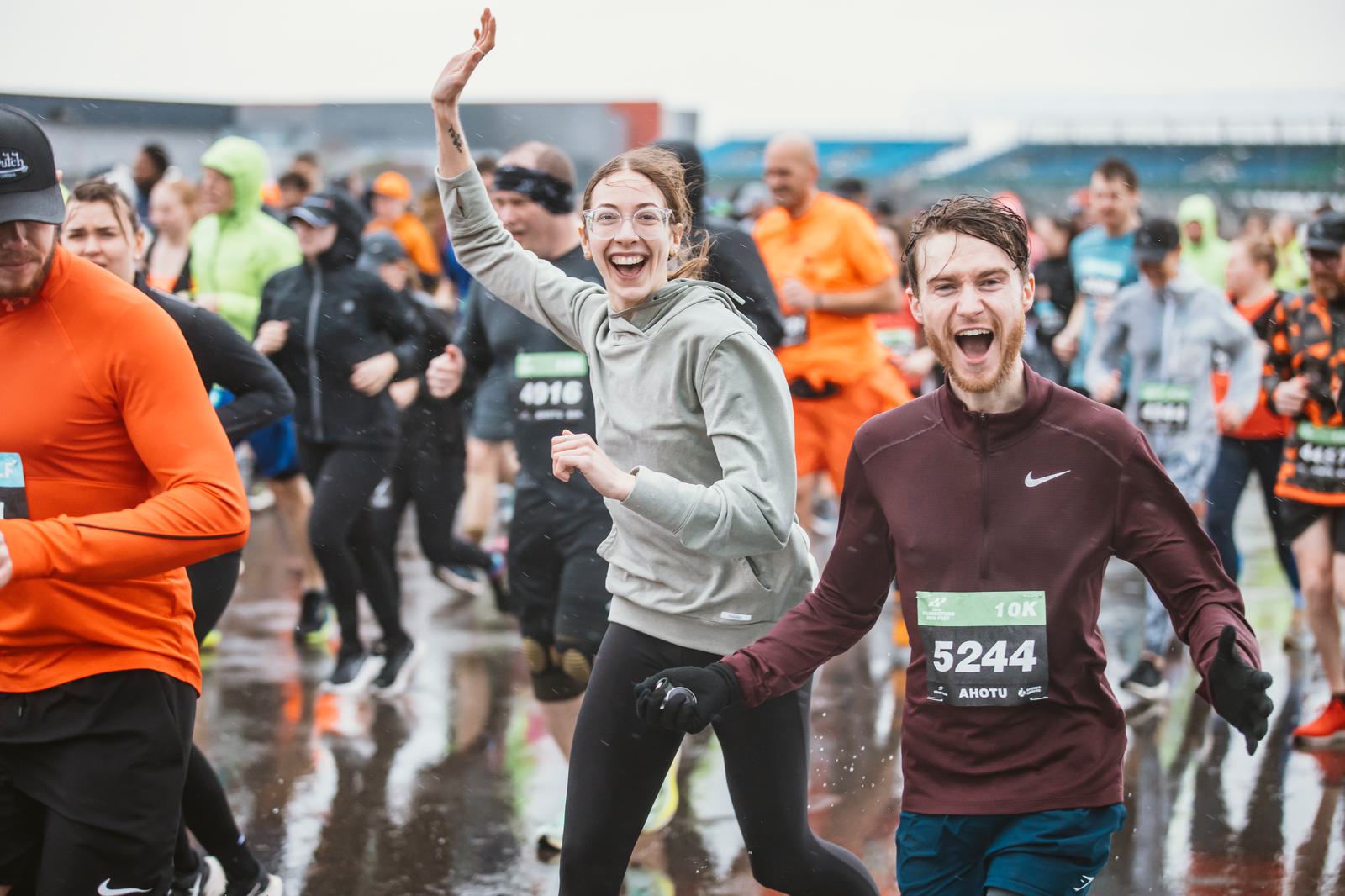Runners participate in a rainy race. A smiling woman waves while a man next to her, wearing a maroon jacket with bib number 5244, grins energetically. The ground is wet, and other runners in colorful attire are visible in the background.