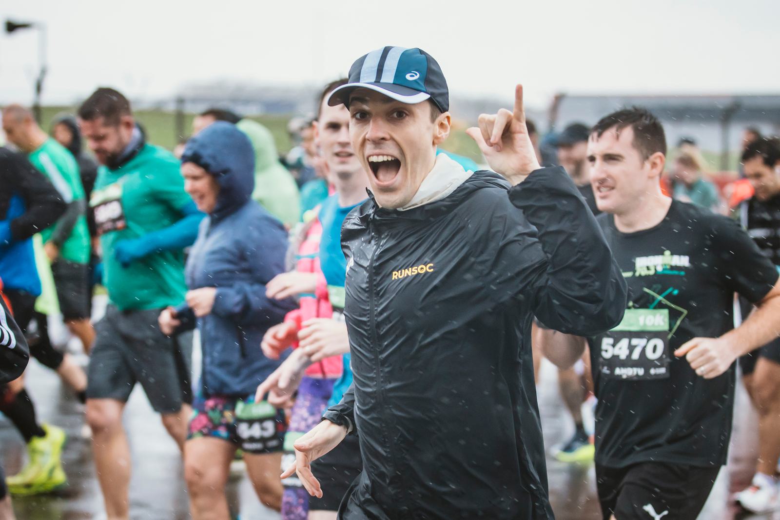 A group of runners participating in a race on a rainy day. The central figure, wearing a black jacket and blue cap, looks excited and is making a gesture with one finger up. Other runners in varied outfits surround them, with water droplets visible.
