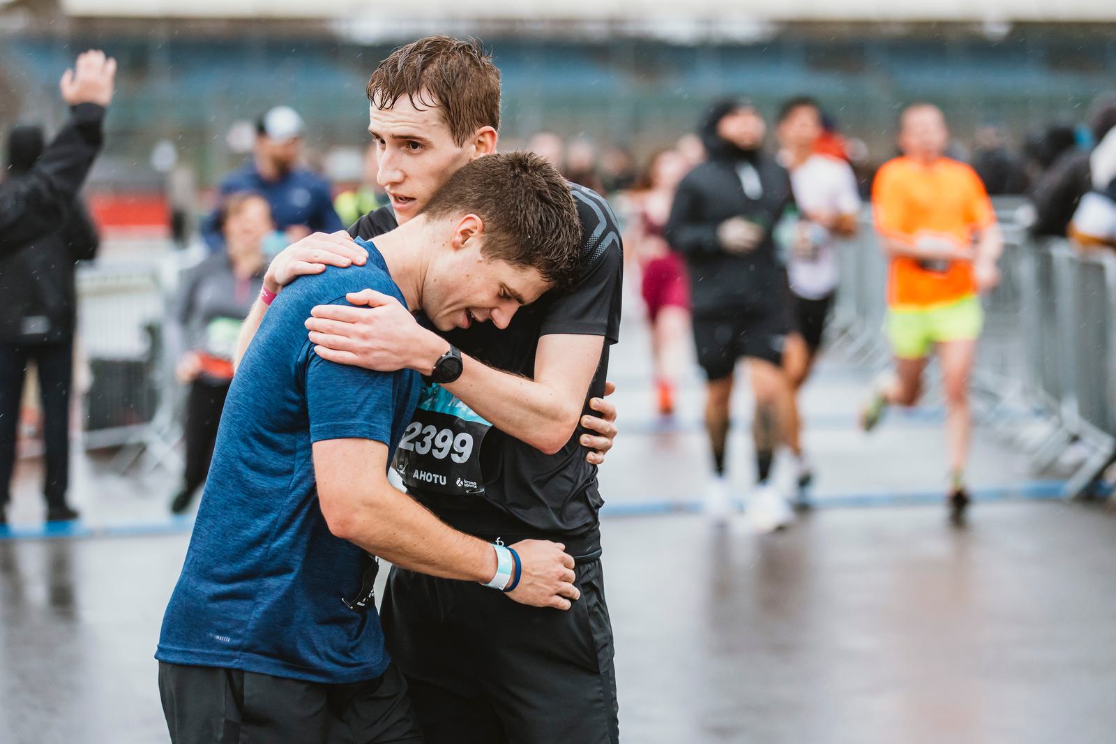 Two men in athletic wear embrace near the finish line of a rainy race. One wears a blue shirt and the other a black shirt, both appearing weary yet supportive. Blurred background shows other runners and a metal barrier.
