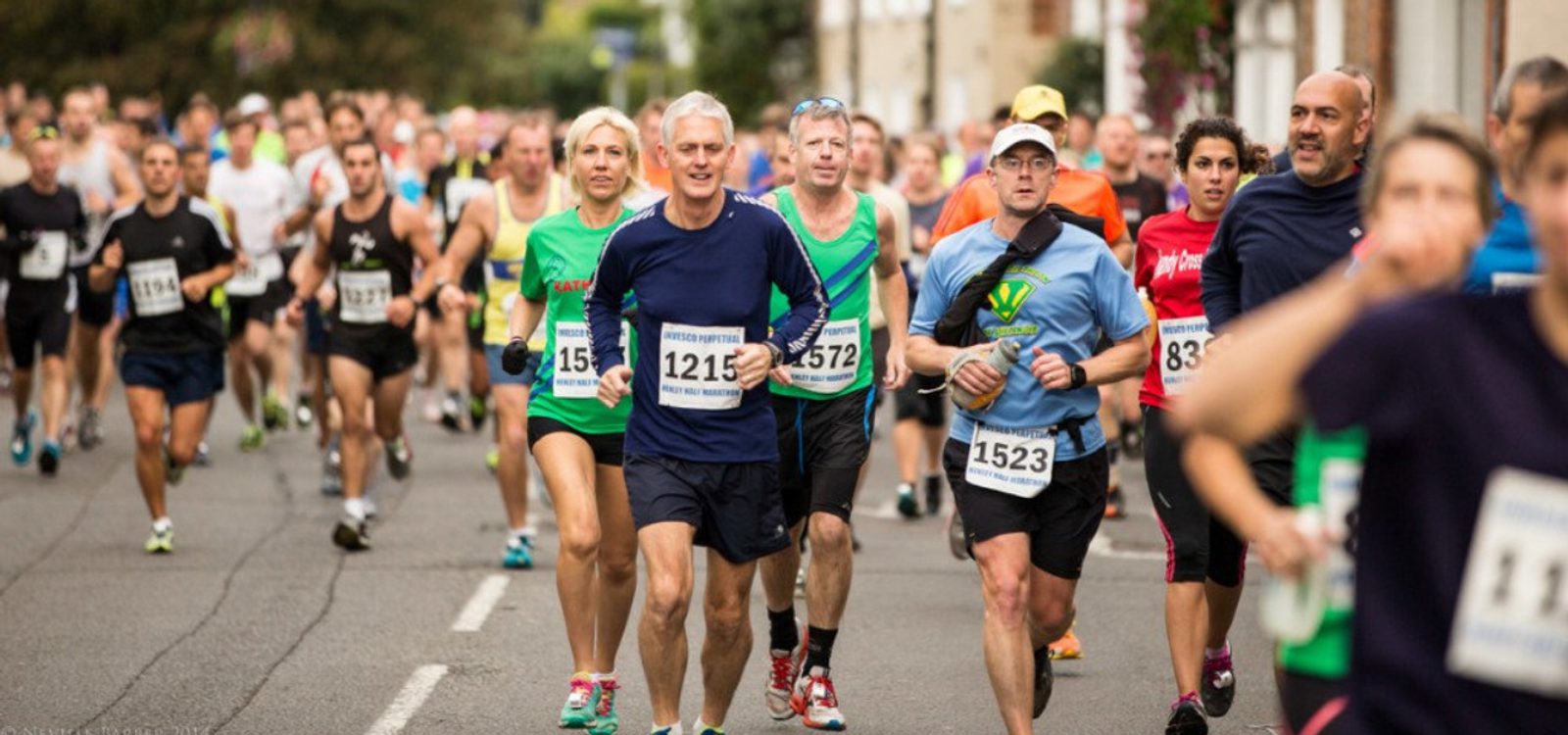 A large group of runners participate in a road race. Men and women of varied ages wear colorful athletic clothing and bib numbers. Some appear focused while others smile. The background shows a tree-lined street with spectators and buildings.