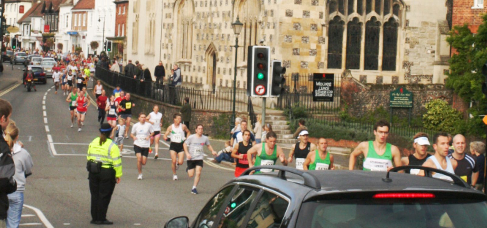 A large group of runners on a street participate in a race near a historic stone building. Spectators watch from the sidewalk. A police officer monitors the event near a traffic light. The scene includes various cars and buildings lining the street.