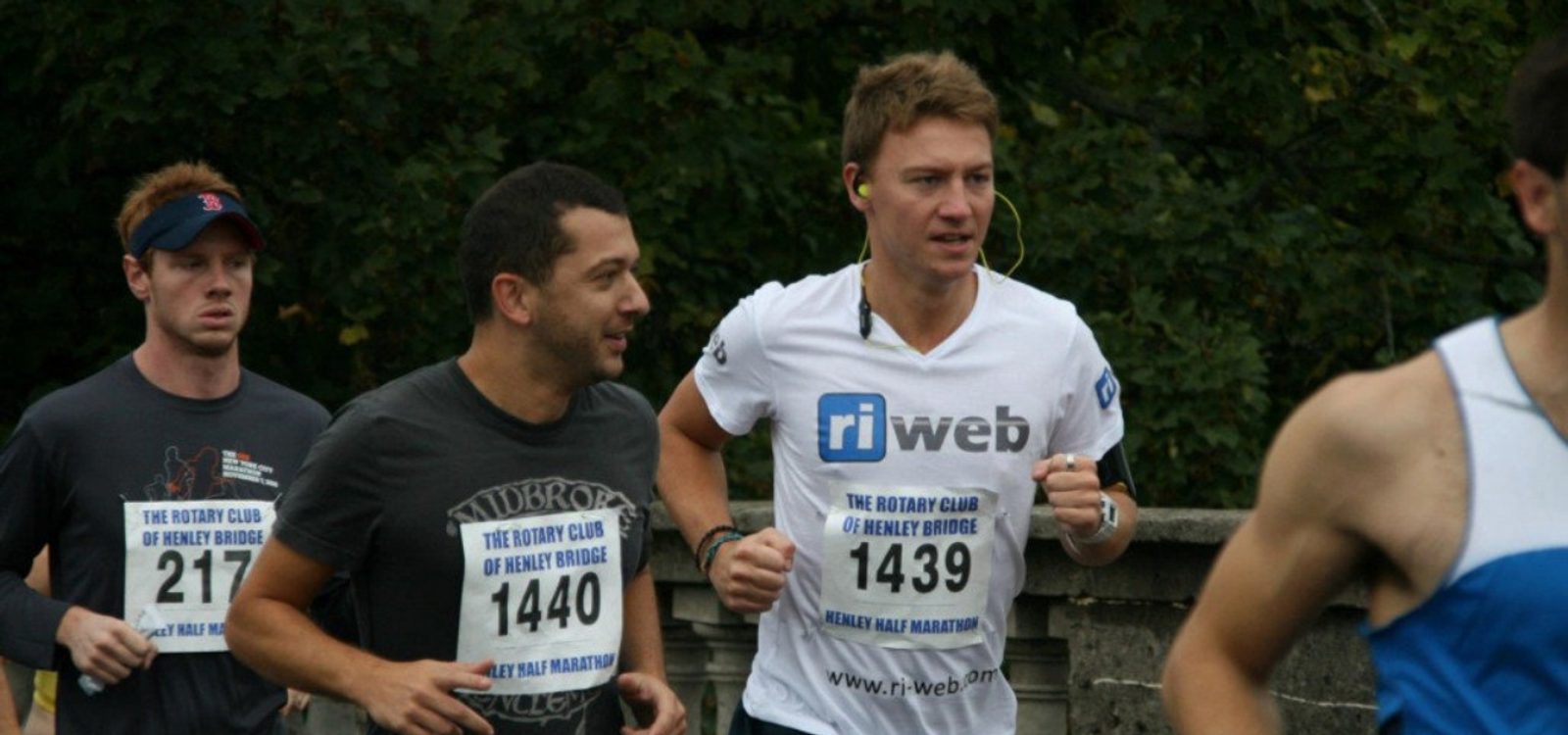 Three men are running in a marathon, all wearing numbered bibs. The man on the right is wearing a t-shirt with the text “ri web.” The background consists of trees and a stone fence. They appear focused and determined as they run.