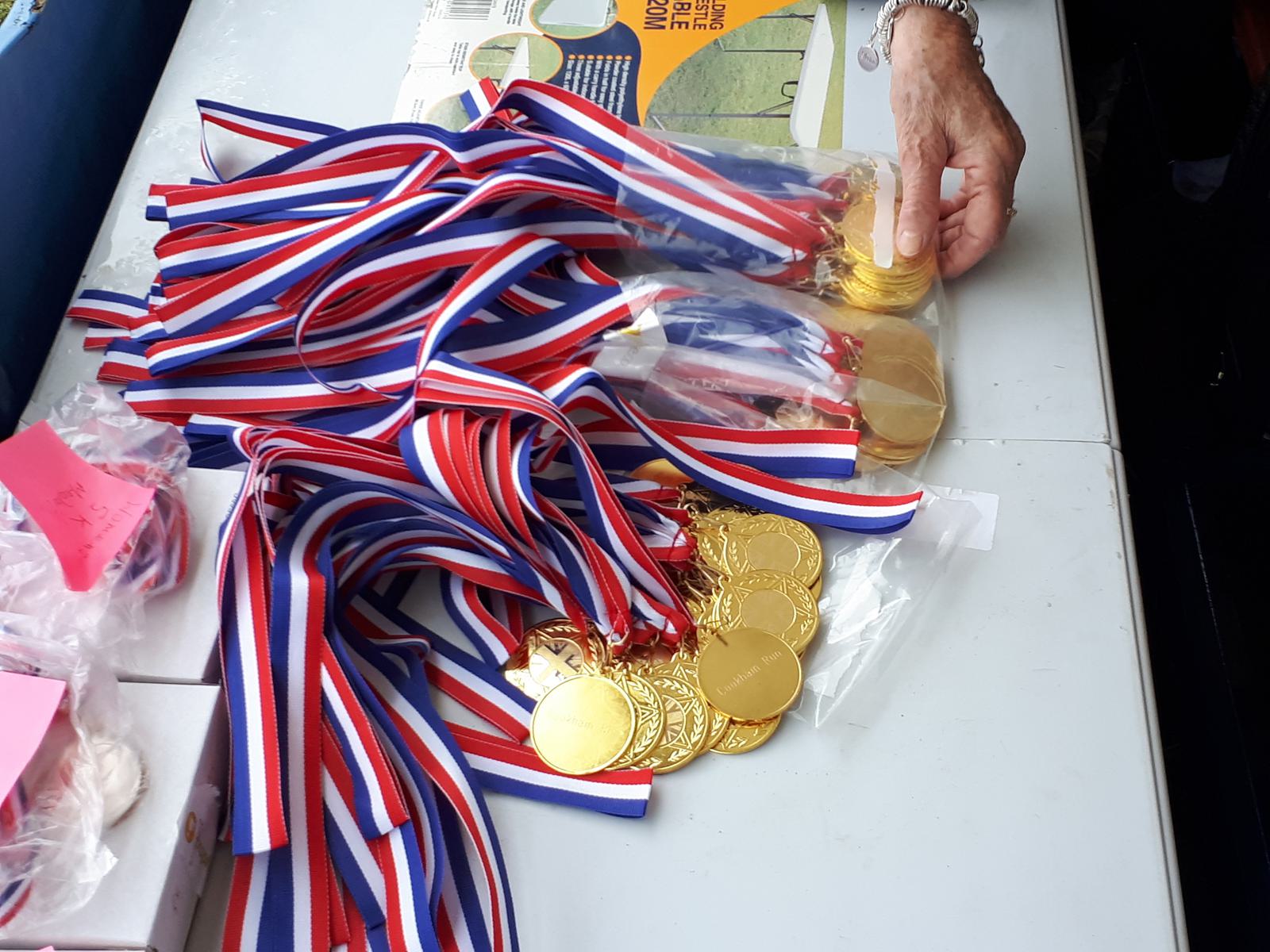 A hand reaches for a stack of gold medals with red, white, and blue ribbons on a table. Several medals are piled up, with colorful ribbons visible. Pink sticky notes and a box are also on the table.