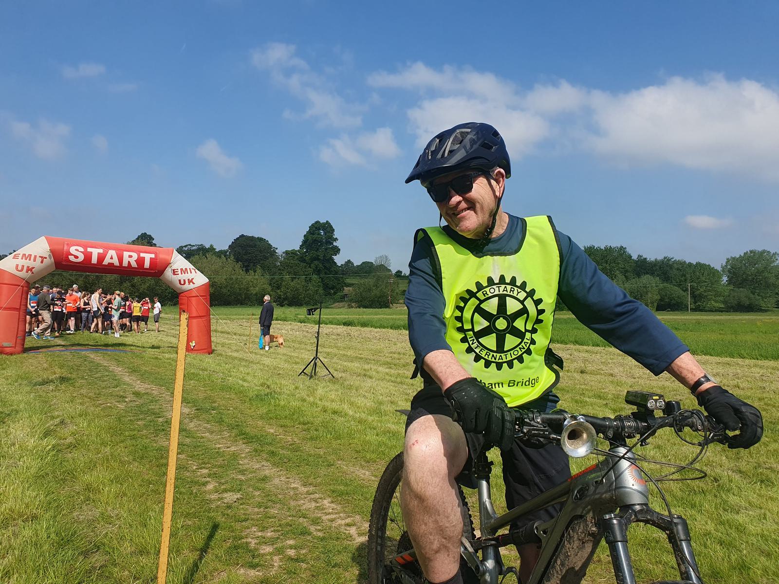 A person wearing a helmet and a neon green vest with a logo rides a bicycle on a grassy path. In the background, a group of people stands near a red "START" archway. The sky is clear and blue.