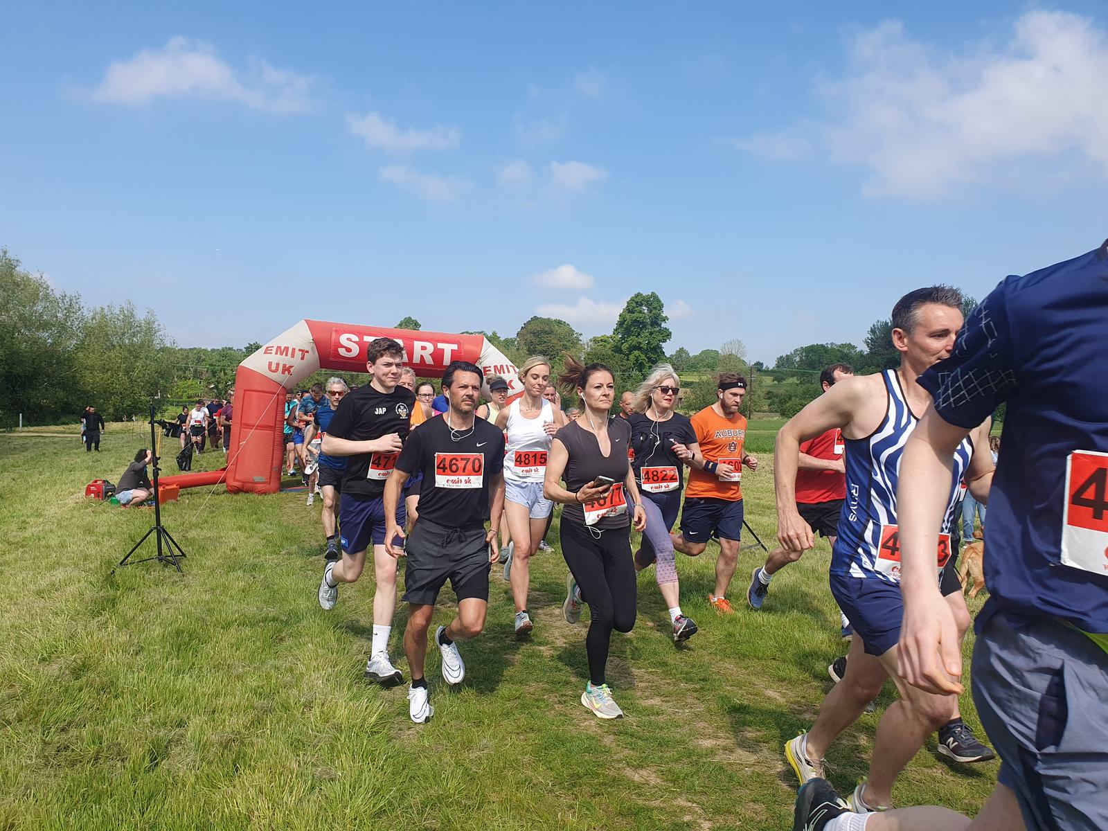 A group of runners starting a race on a grassy field under a blue sky. They are wearing athletic gear and numbered bibs, with a red starting arch in the background. Trees and spectators can be seen in the distance.