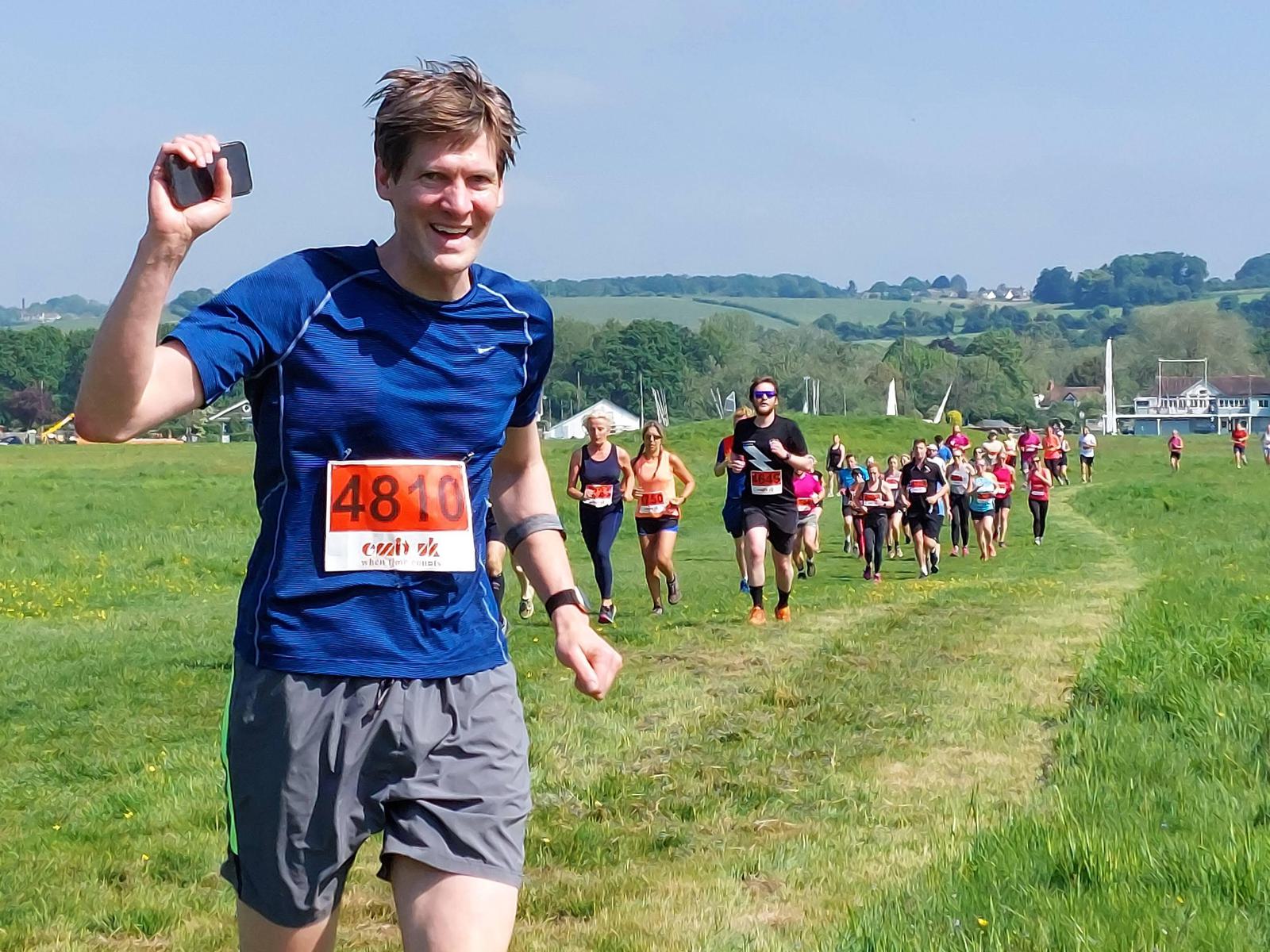 A smiling runner in a blue shirt and gray shorts holds up a phone while participating in a race on a grassy field. He wears a race number bib, and more participants run behind him under a clear blue sky.