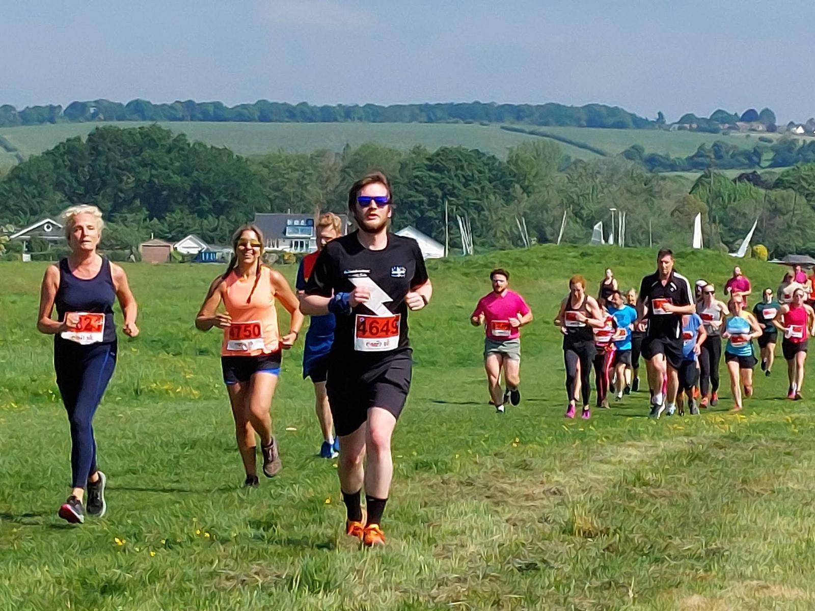 A group of runners participating in a race on a grassy field under clear blue skies. The front runners wear numbered bibs. A scenic landscape with rolling hills and trees is visible in the background.