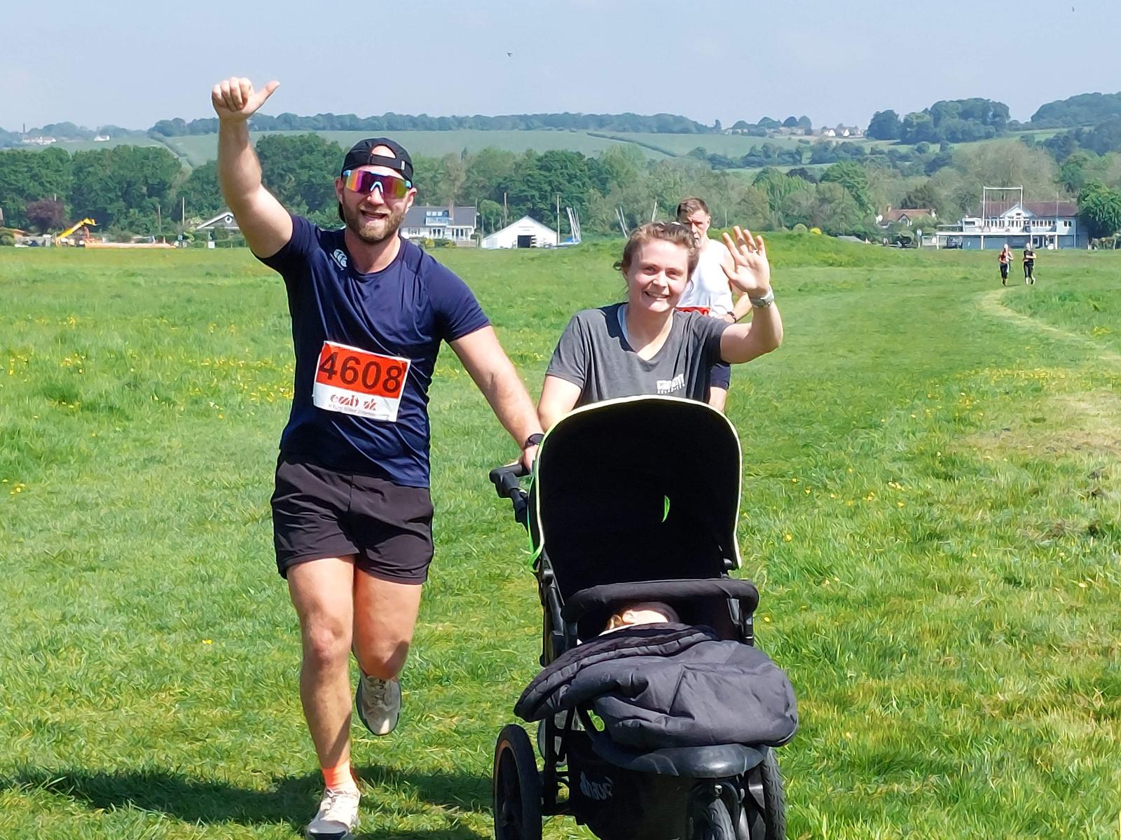 A man and a woman jog on a grass path, smiling and waving. The man wears a "4608" race bib, sunglasses, a cap, a navy shirt, and black shorts. The woman pushes a stroller, wearing a gray shirt and black shorts. Lush hills and buildings are in the background.