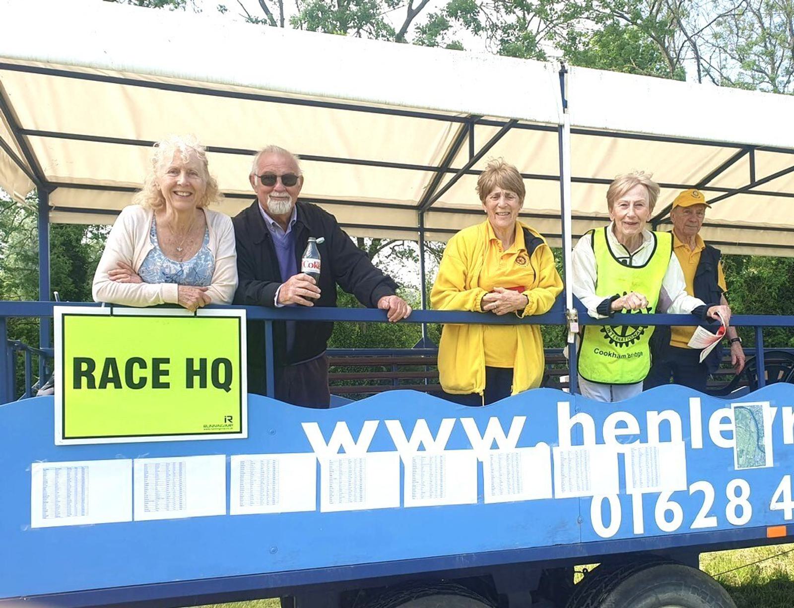 Five people stand on a platform under a canopy at a race event. A sign reads "RACE HQ." They appear to be smiling and some are wearing yellow jackets. Trees are visible in the background.