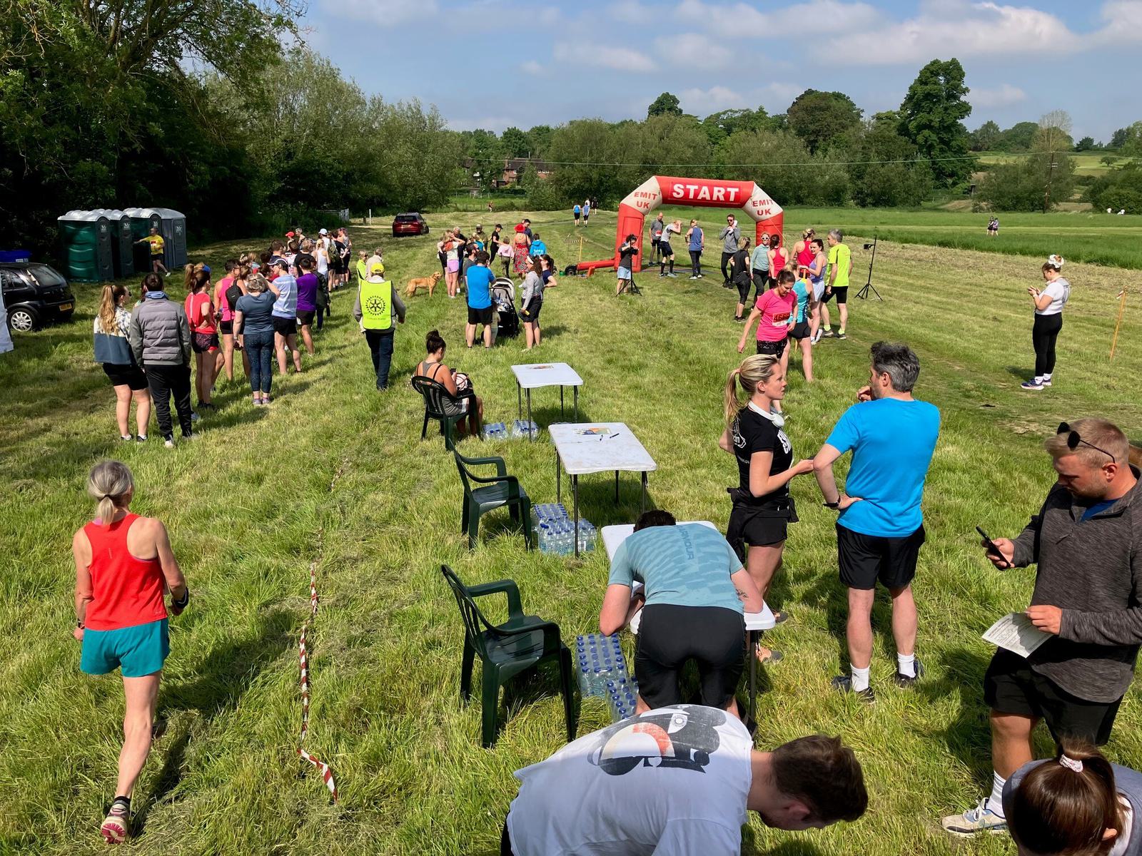 A group of runners gather on a grassy field near a red "START" arch. Tables are set up with chairs. People are preparing for a race, stretching, and chatting in casual athletic wear. The sky is partly cloudy, and trees are in the background.