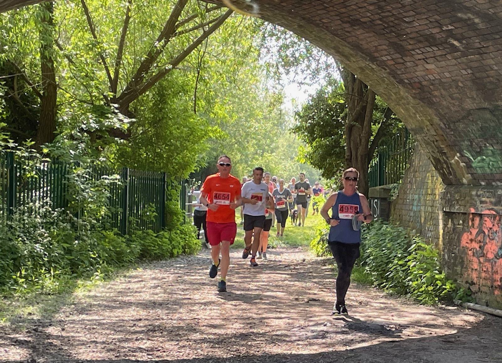 Runners participating in a race under a brick bridge on a sunny day, surrounded by greenery. The path is shaded by trees, and sunlight filters through leaves, casting patterns on the ground. The lead runner wears a red shirt and sunglasses.