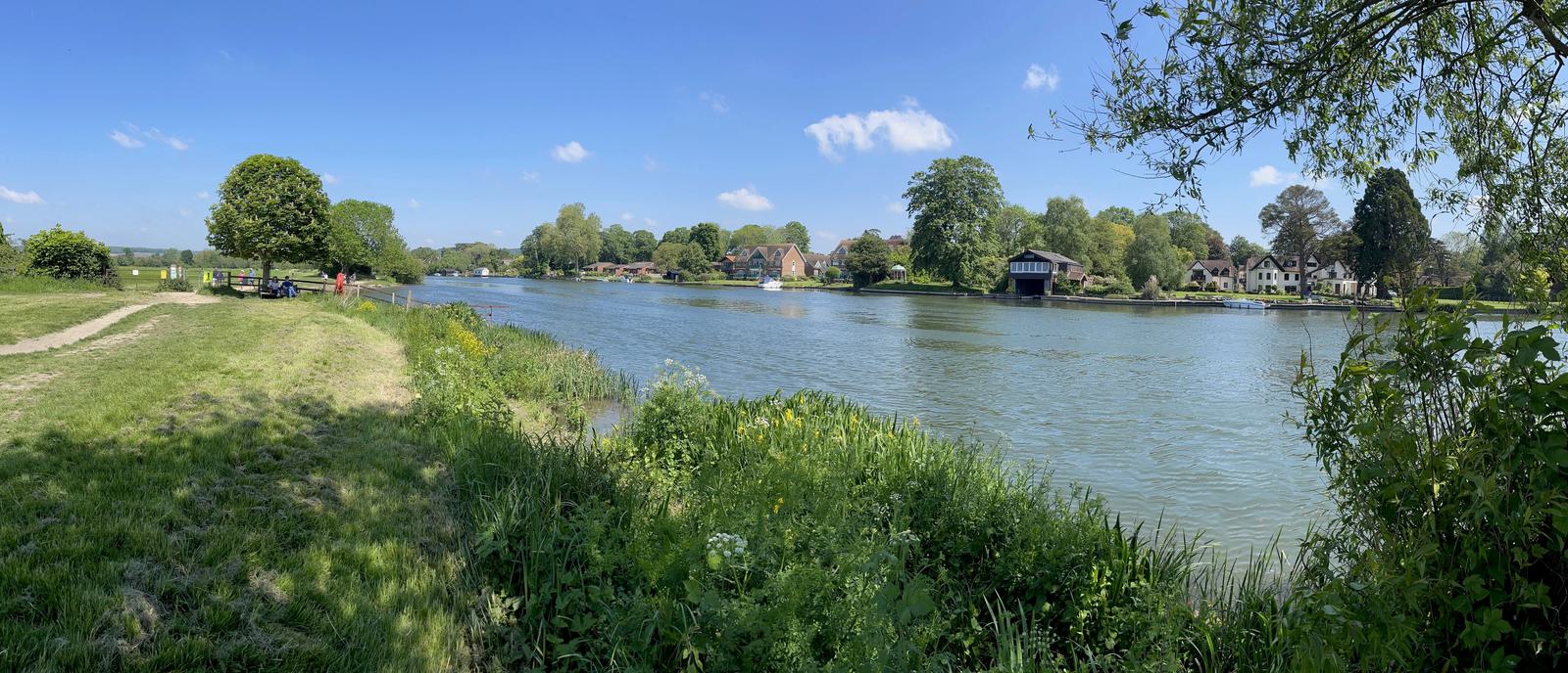 Panoramic view of a wide river with lush green banks and trees. Houses are visible along the opposite shore under a bright blue sky with scattered clouds. A path and picnic table are in the foreground to the left.