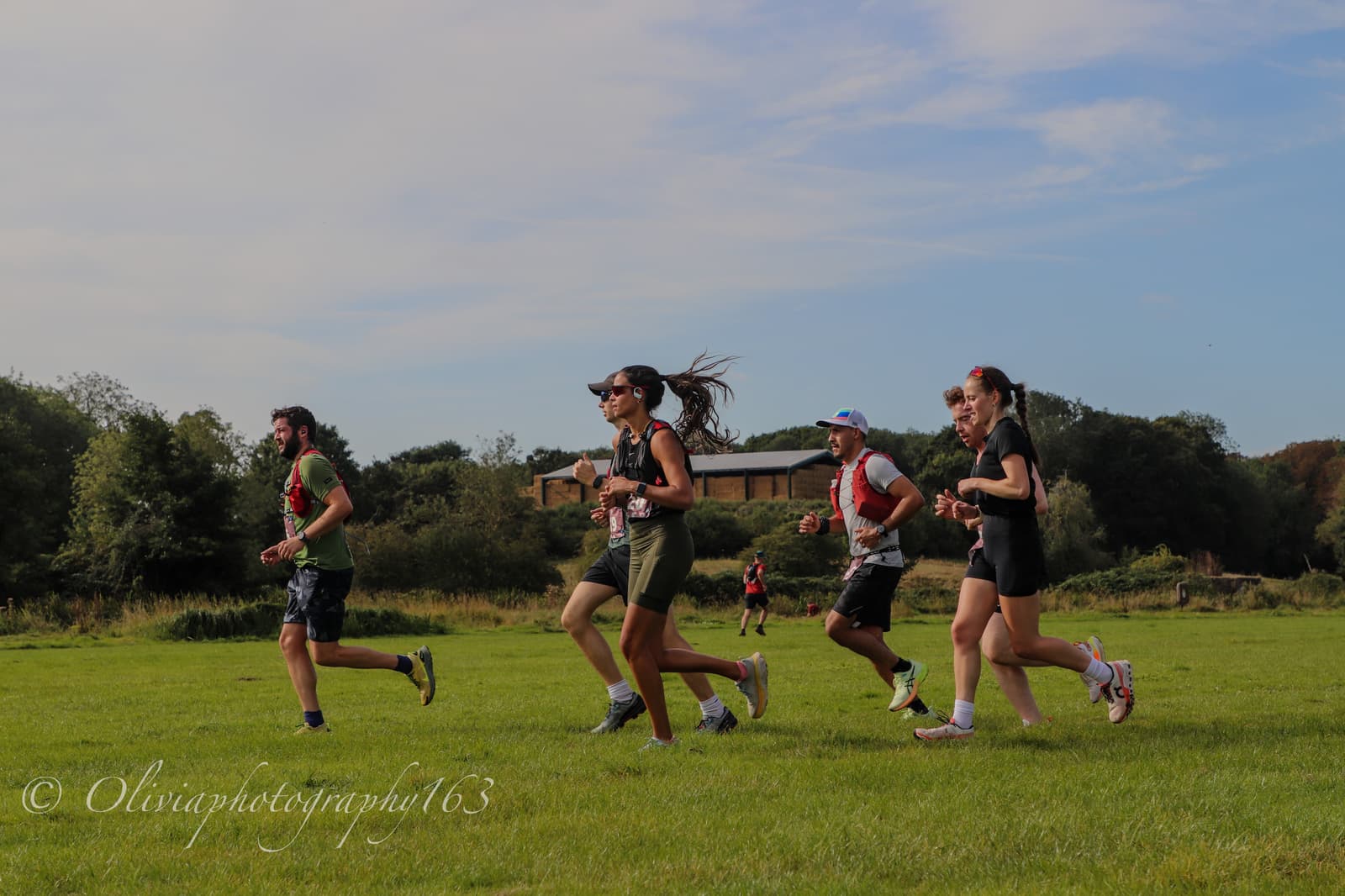 Five people are running outdoors on green grass, with trees and houses in the background under a blue sky. They appear to be participating in a race or organized run.