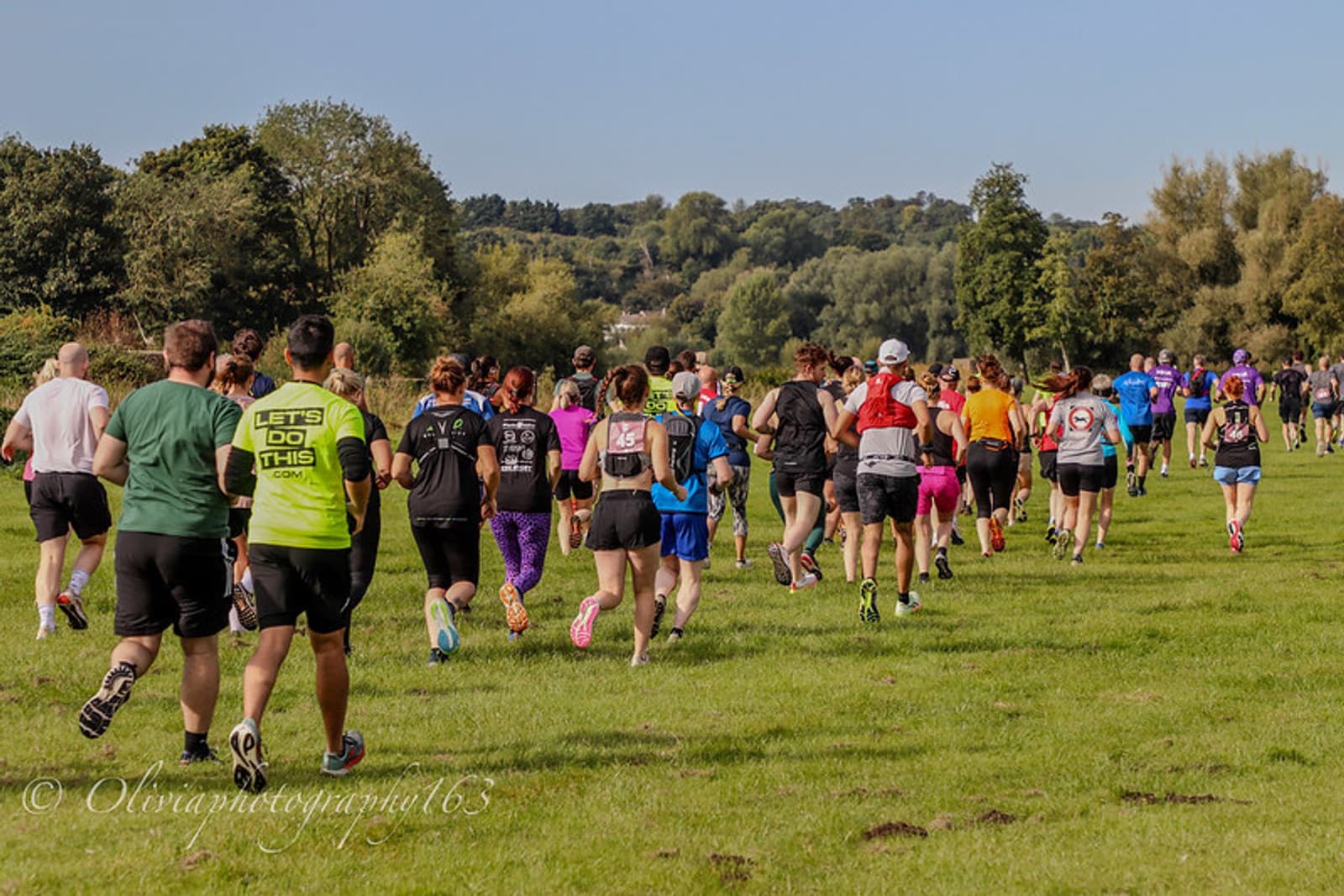 A group of people running together outdoors on a grassy field under a clear, sunny sky, surrounded by green trees and distant hills. Most runners wear colorful athletic clothes.