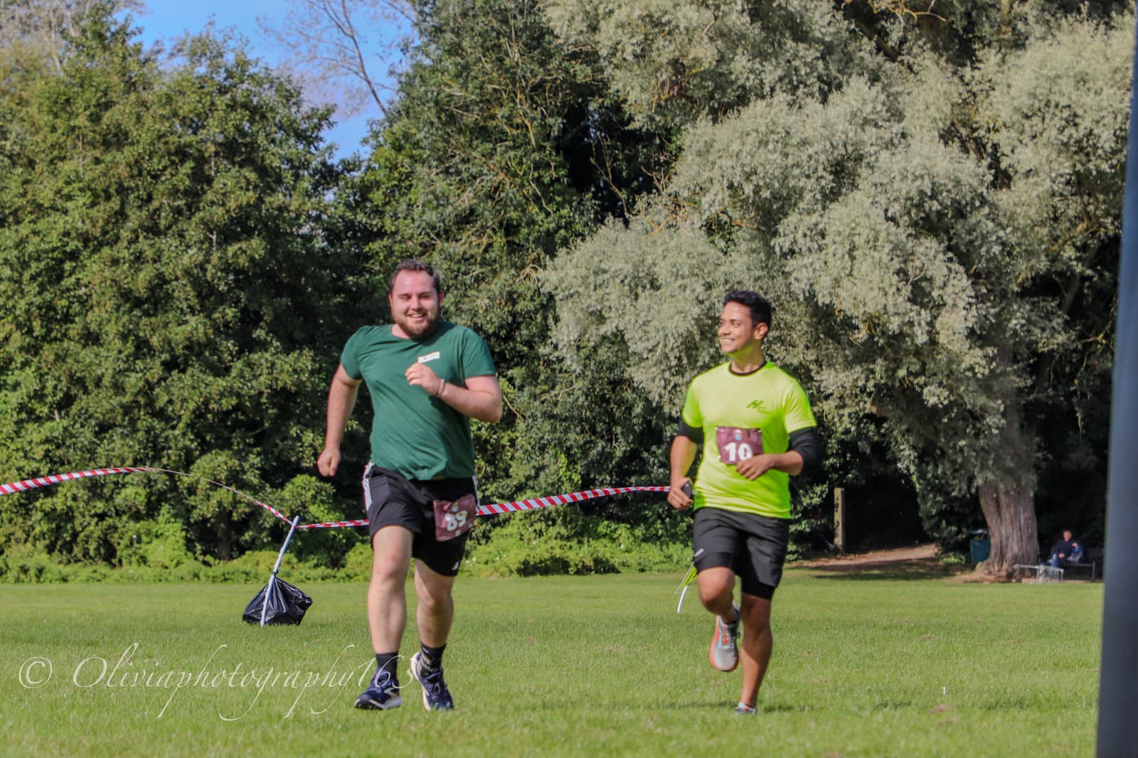 Two men wearing race bibs are running toward a finish line on a grassy field, surrounded by trees. One man in a green shirt is smiling as he runs ahead of the other, who is wearing a bright yellow shirt.