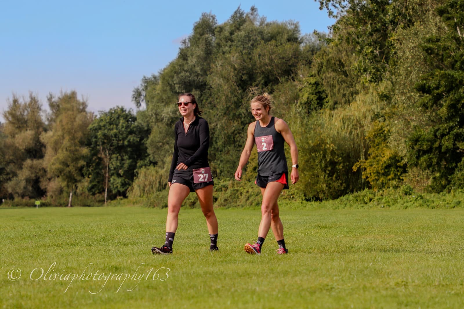 Two women wearing running attire and race bibs walk on a grassy field, smiling, with trees and blue sky in the background.