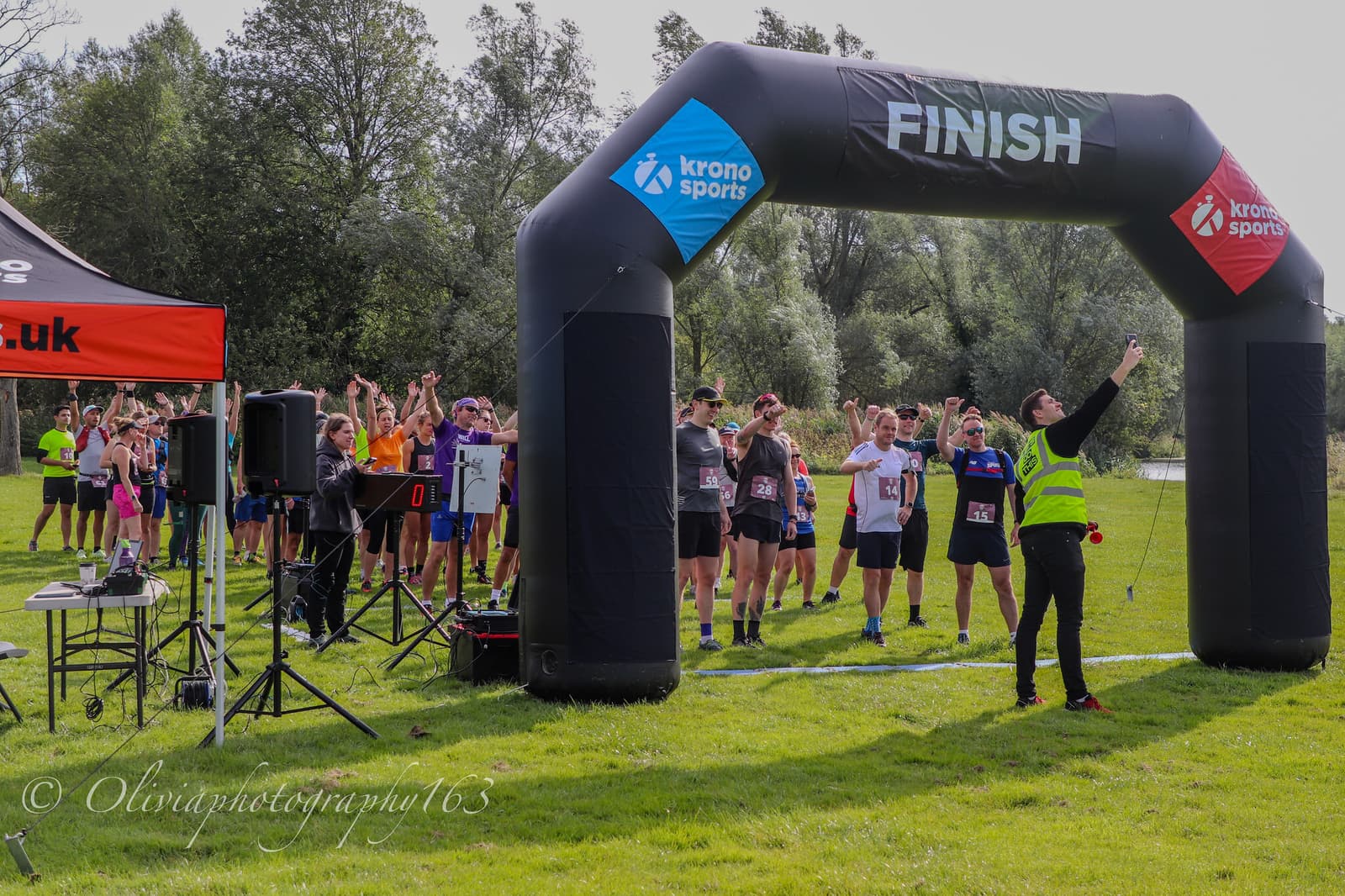 A race official in a yellow vest takes a selfie under a black inflatable finish arch, while a group of runners stand behind the start line, some with arms raised, on a grassy field with trees in the background.
