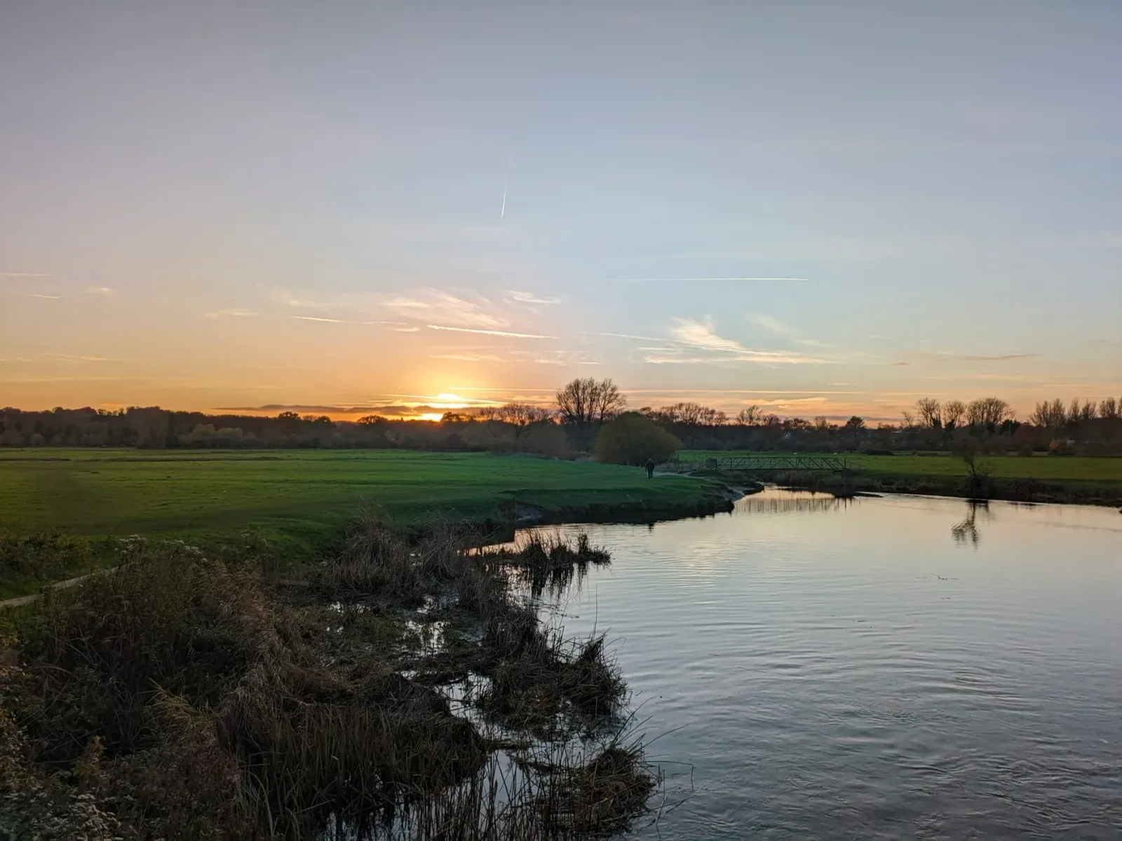 A serene landscape showcasing a picturesque sunset over a calm river. The water reflects the warm, golden hues of the sky, while the surrounding green fields and distant trees create a tranquil atmosphere, capturing the beauty of nature as day transitions to night.