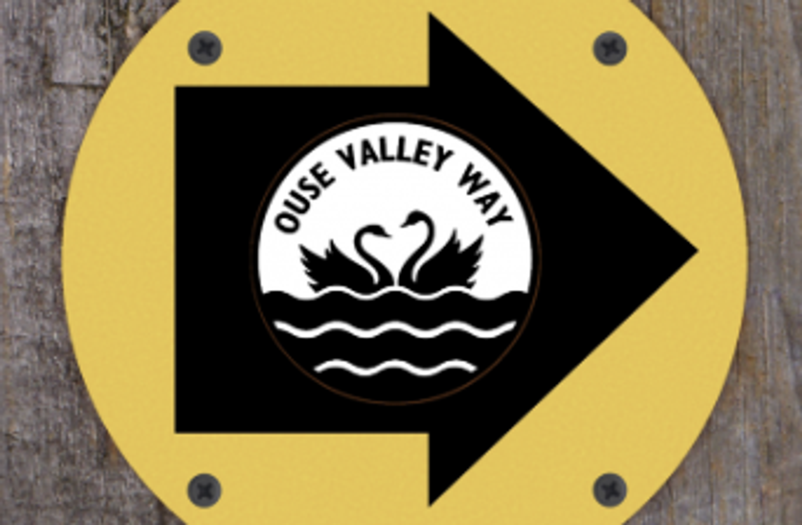 A yellow circular sign with a black rightward arrow and an emblem in the center. The emblem features two swans facing each other with the text "OUSE VALLEY WAY" above them and wavy lines below representing water. The sign is mounted on a wooden surface.
