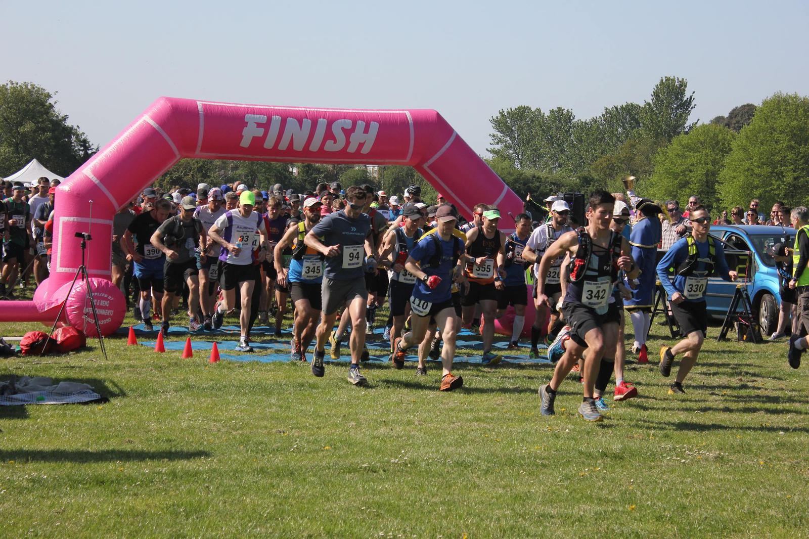 A group of runners starts a race, passing under a large pink arch marked "FINISH." They are outdoors on a grassy field, with trees in the background and spectators watching. The runners wear numbered bibs and various athletic gear.