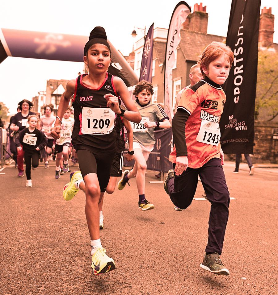 Children wearing race numbers are running in a street race. An inflated arch with "SPORTS" is visible in the background. The atmosphere is energetic, with spectators lined along the sides. The sky is overcast.