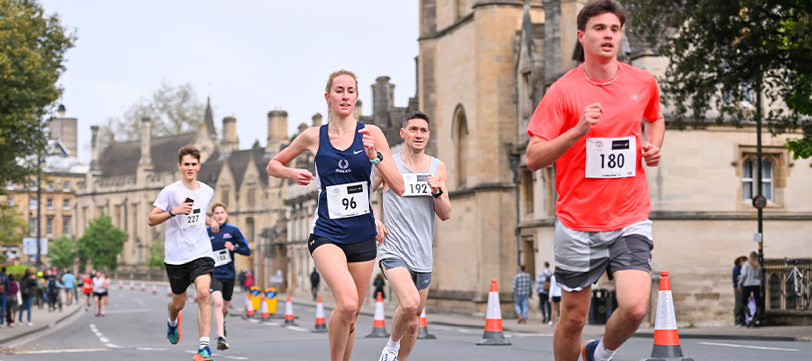 A group of runners participates in a road race. They are on a street lined with historic buildings. Traffic cones separate the runners from spectators. The sky is overcast, and trees are visible in the background.