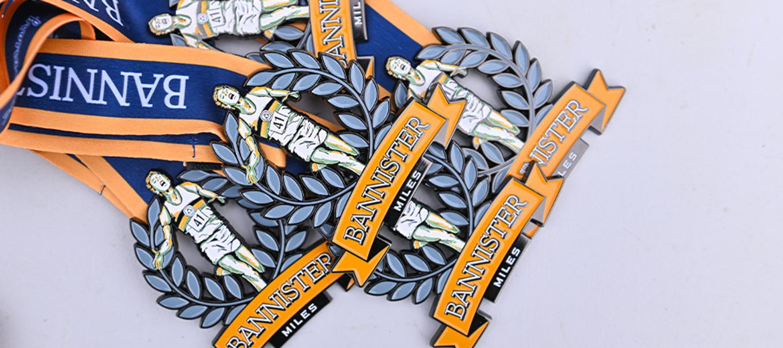 A pile of Bannister Miles medals with blue and orange ribbons. Each medal features an illustration of a runner and the words "Bannister Miles" on an arrow-shaped banner.