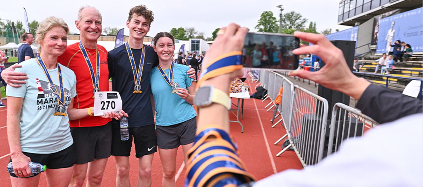 Four people, wearing running gear and medals, smile for a photo on a running track. A person in the foreground holds a smartphone to capture the moment. The background includes bleachers and event banners.