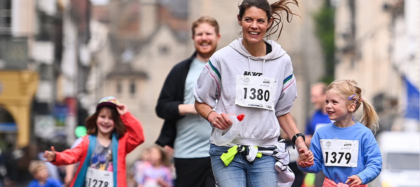A woman and two children, all wearing race bibs numbered 1380, 1379, and 1278, run joyfully in a street race. They are dressed in casual athletic wear, and a smiling man appears in the background. The scene is lively and cheerful.