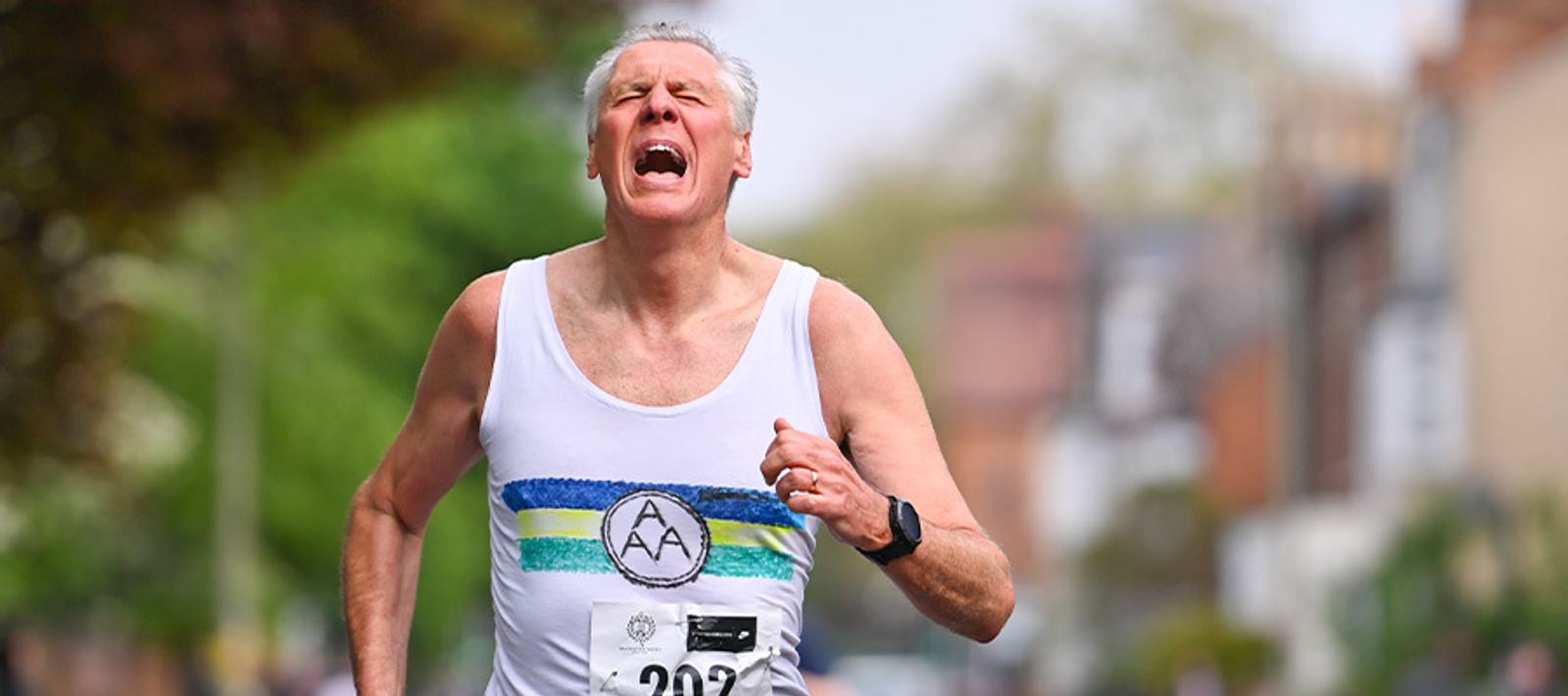 An older man is running in a race, wearing a white tank top with a purple and green stripe and displaying a race number. His expression shows exertion. The background is slightly blurred with greenery and buildings.