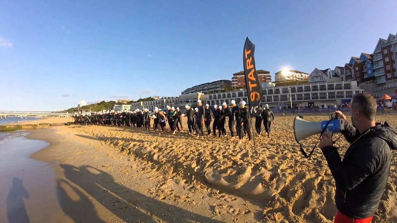 A group of triathletes, wearing wetsuits and swim caps, walk toward the starting line of a beach triathlon. A man holds a large megaphone, and a "START" banner marks the beginning. People are gathered in the background near buildings on a sunny day.