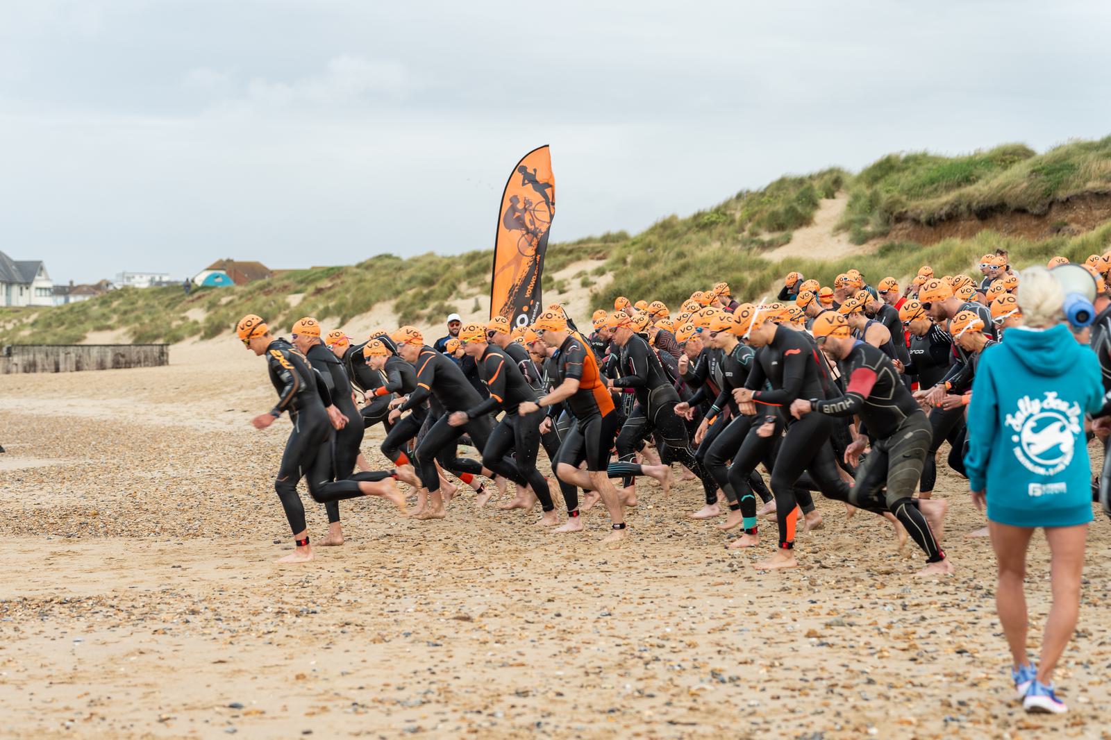 A group of athletes wearing wetsuits and orange swim caps are running along a pebble beach, preparing for a water-based race. A flag with a graphic stands upright in the background. A person in a blue shirt observes the scene.