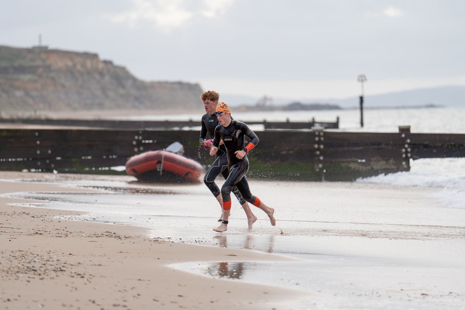 Two athletes wearing wetsuits and swim caps run on a sandy beach. An inflatable boat and rocky coastline are visible in the background, with calm sea waves approaching the shore. The sky is cloudy.