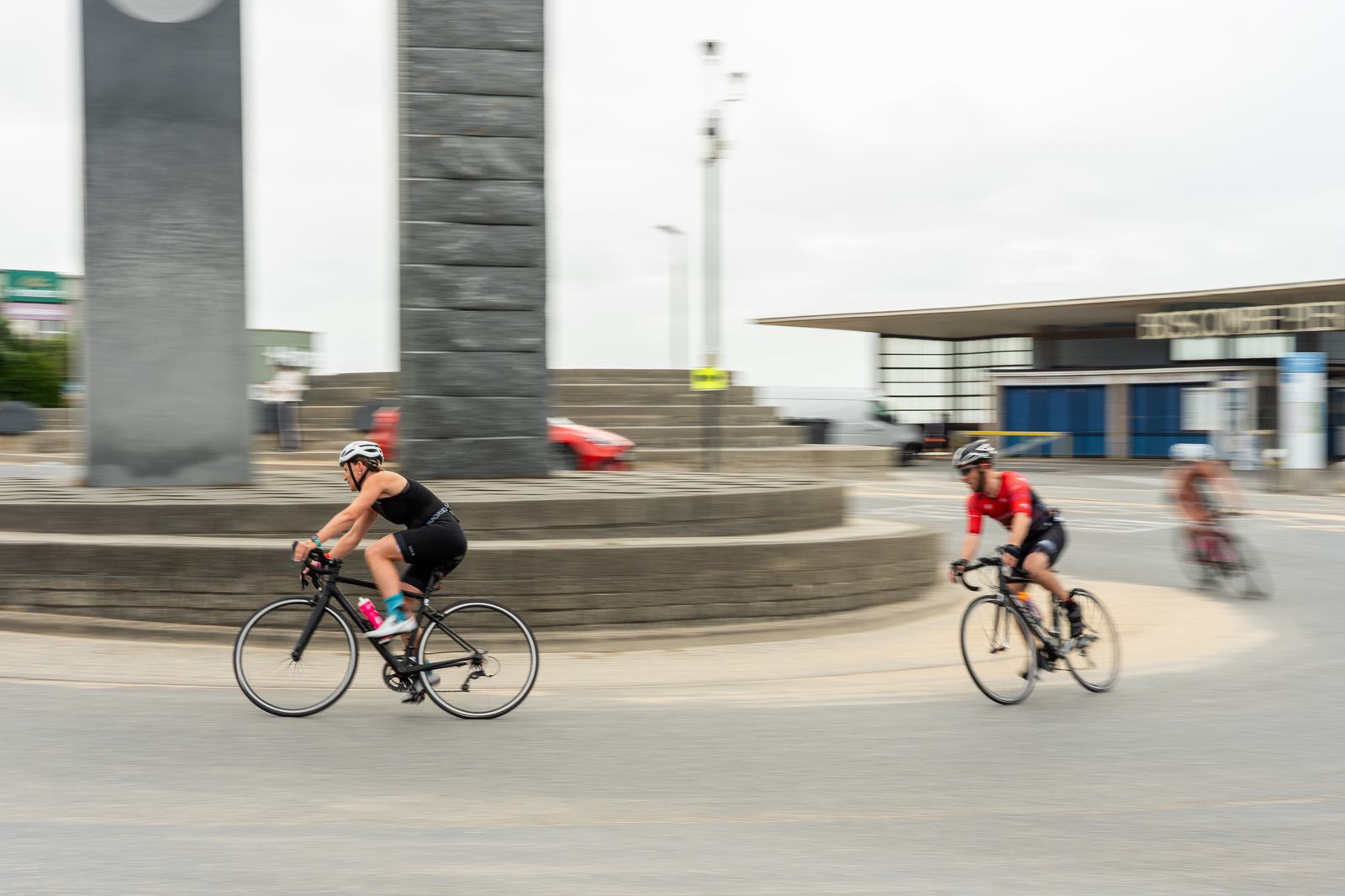 Cyclists in motion ride around a circular stone monument with one leading in a black outfit and the other in red. A blurred third cyclist follows in the background. The cloudy sky enhances the sense of movement.