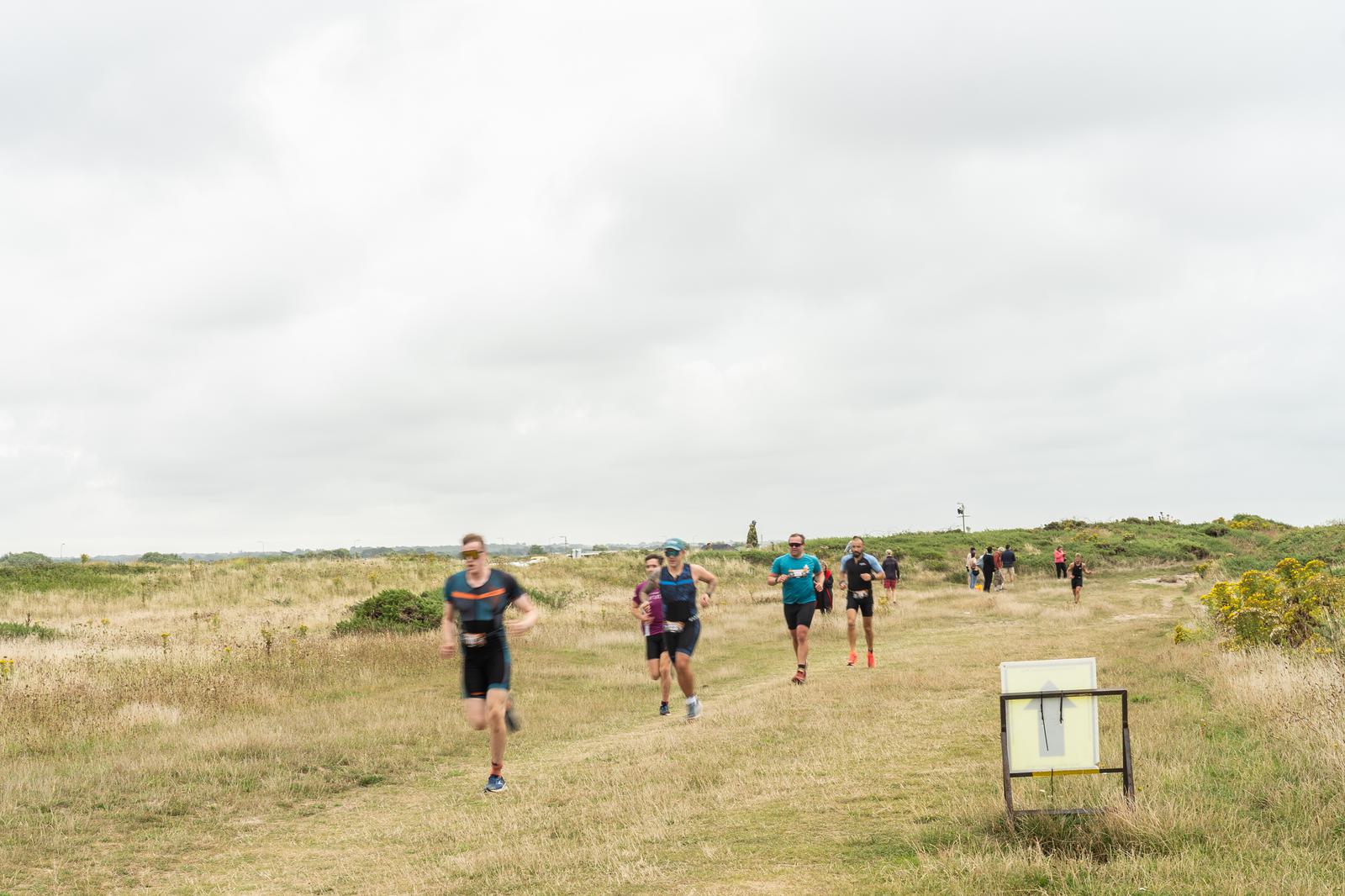 Runners participating in a race on a grassy trail, surrounded by open fields under a cloudy sky. The path is marked by a sign in the foreground.