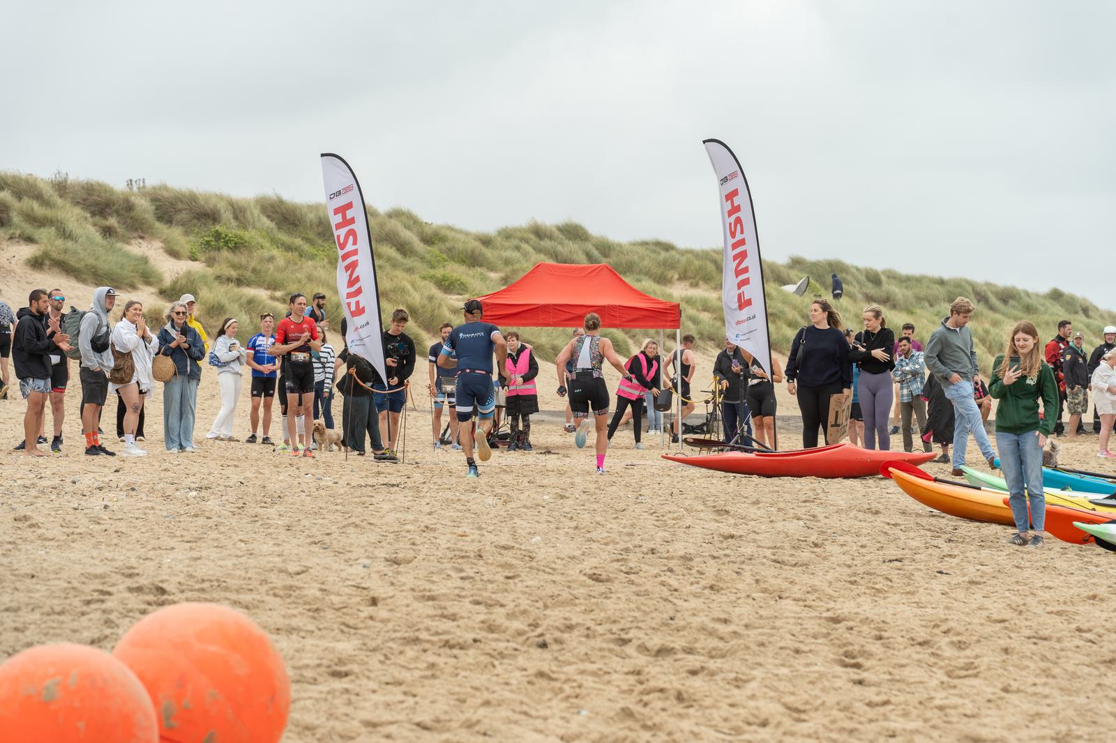 Participants in a beach race sprint towards the finish line, marked by two tall flags. Spectators stand nearby, while colorful kayaks and large orange buoys are visible on the sand. A red tent is set up in the background under a cloudy sky.