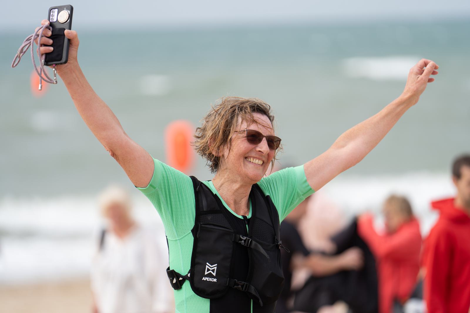 A person wearing sunglasses and a wetsuit vest joyfully raises their arms on a beach. They are holding a small device in one hand. The ocean is in the background, with a partly cloudy sky. People in various outfits are visible in the distance.