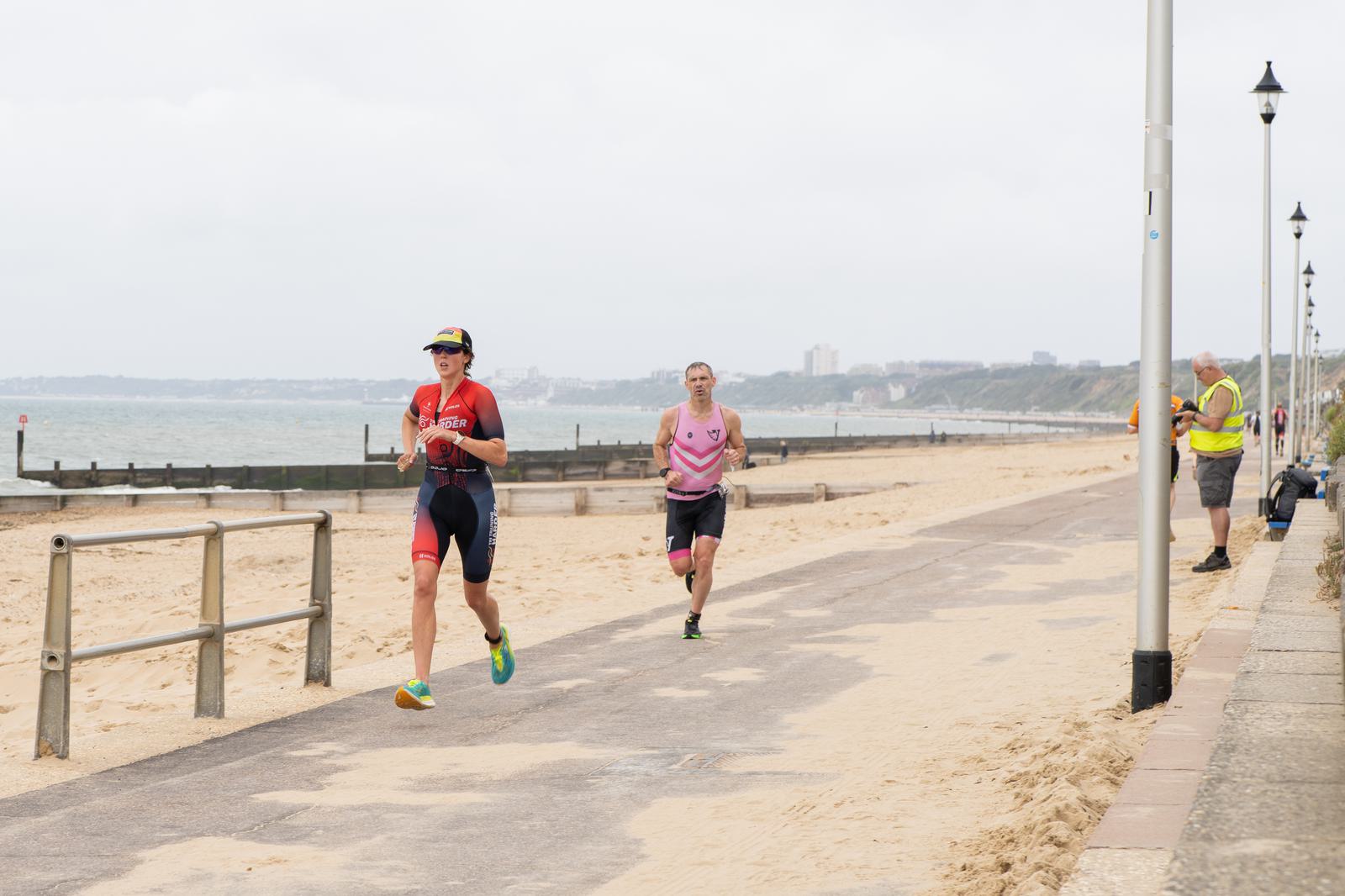 Two athletes running on a coastal path during a triathlon. The athlete in front wears a black and red suit, and the one behind wears pink and gray. The path is lined with lampposts and the beach is on the left. A cloudy sky is above.