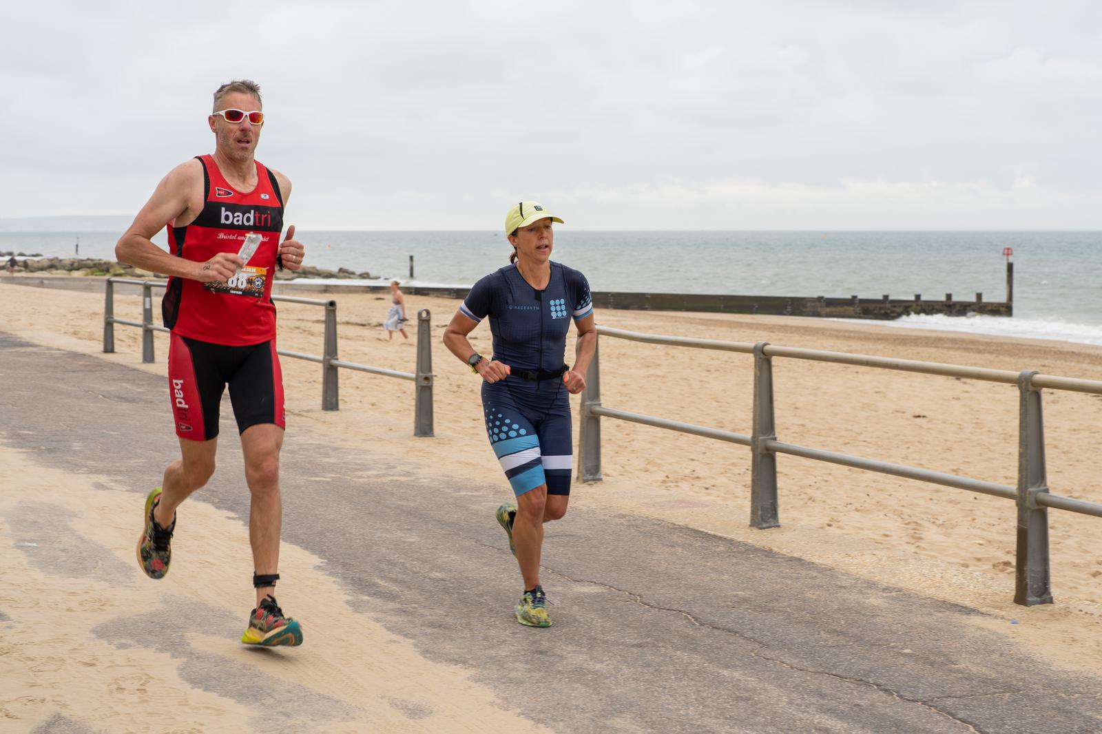 Two people in athletic gear are running on a seaside path. The man on the left wears a red outfit, sunglasses, and a race bib. The smaller person on the right wears a dark blue outfit and a yellow cap. The ocean is visible in the background.