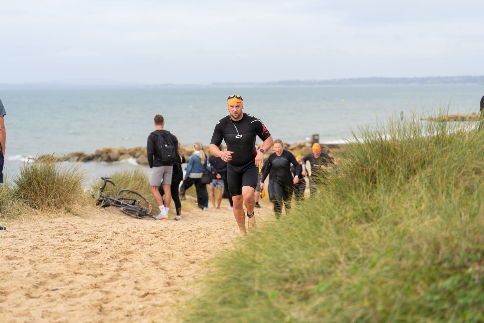 A person in a wetsuit and yellow swim cap runs up a sandy path from the beach, followed by others in similar attire. In the background, people walk along the shore near the ocean. Waves gently crash, and the sky is overcast.