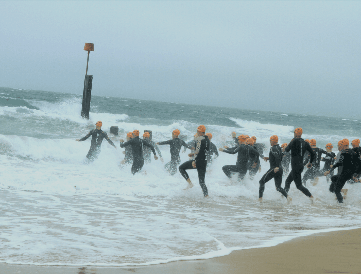 A group of triathletes in black wetsuits and orange swim caps run into the ocean from a sandy beach, preparing to start the swim portion of a triathlon. The sea is wavy and a wooden post with a red navigational marker is visible in the water. The sky is overcast.