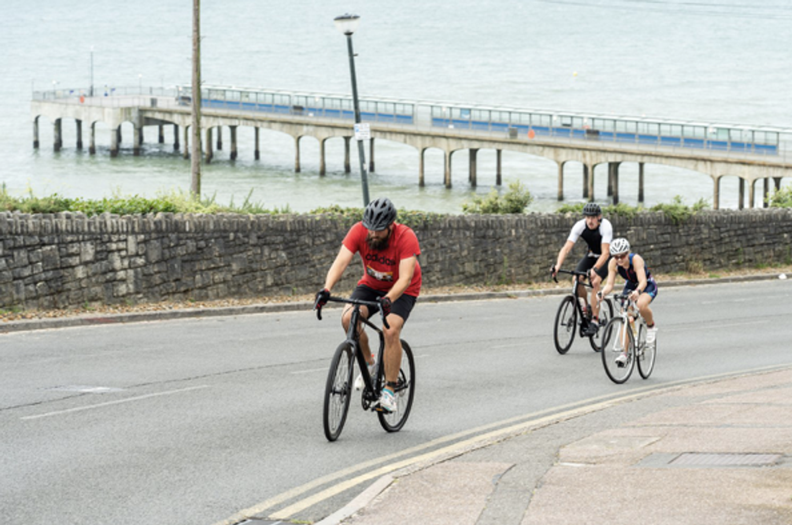 Three cyclists riding uphill on a road near the coast. A stone wall and a pier extending into the water are visible in the background. The cyclists wear helmets, and one is in a red shirt. The scene suggests a pleasant day for outdoor exercise.
