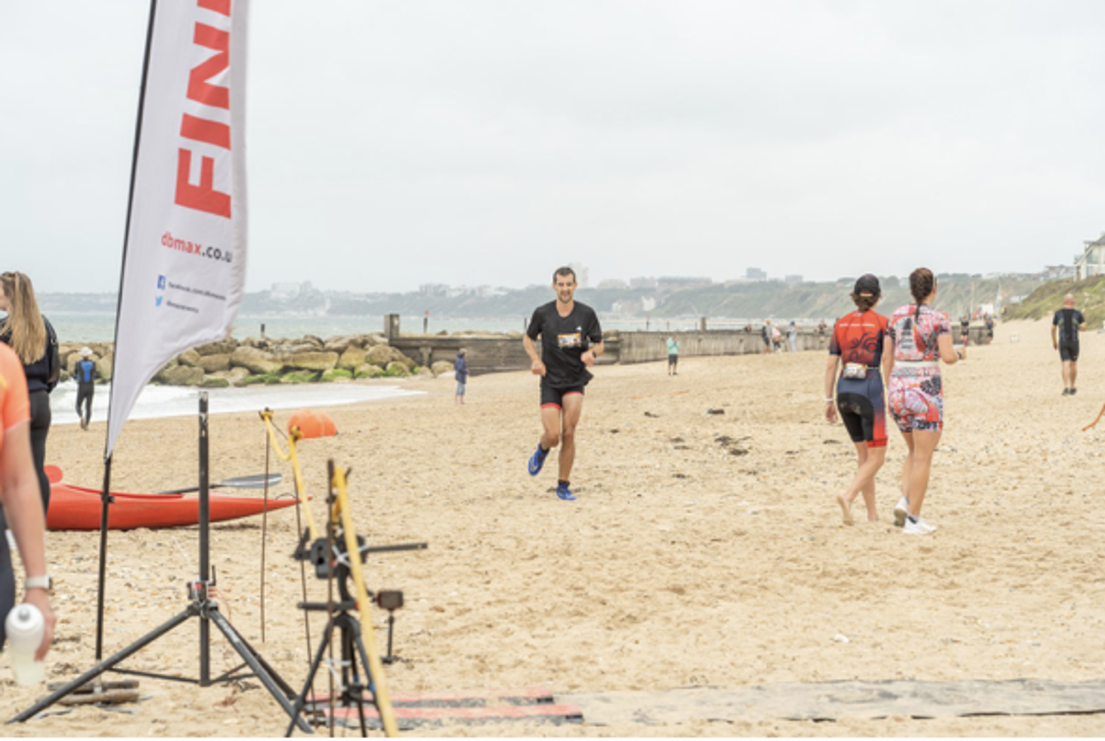A man runs on a sandy beach toward a finish line marked by a flag. Other participants and a few onlookers are present. The sky is overcast, and the shoreline is visible in the background.