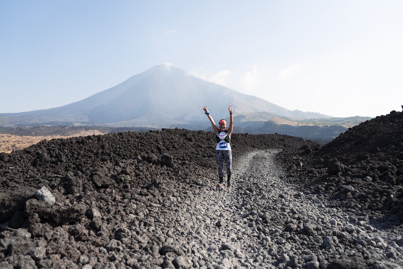 A person is running on a rocky trail with their arms raised in celebration against the backdrop of a large volcano. The landscape around them is formed from dark volcanic rocks, creating a rugged terrain. The sky is clear with a faint haze near the volcanoβs peak.