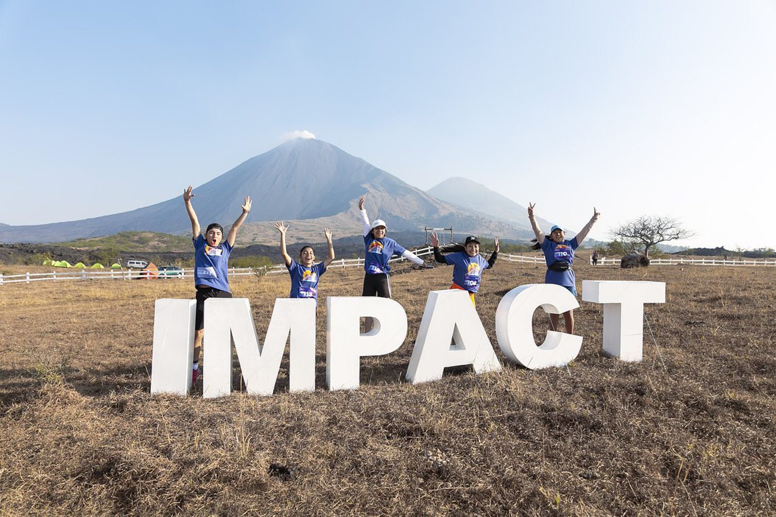 A group of five people wearing blue shirts stand behind large, white letters spelling "IMPACT" in an open field. They are all jumping with arms raised. In the background, there are white fences, tents, and a mountain under a clear blue sky.