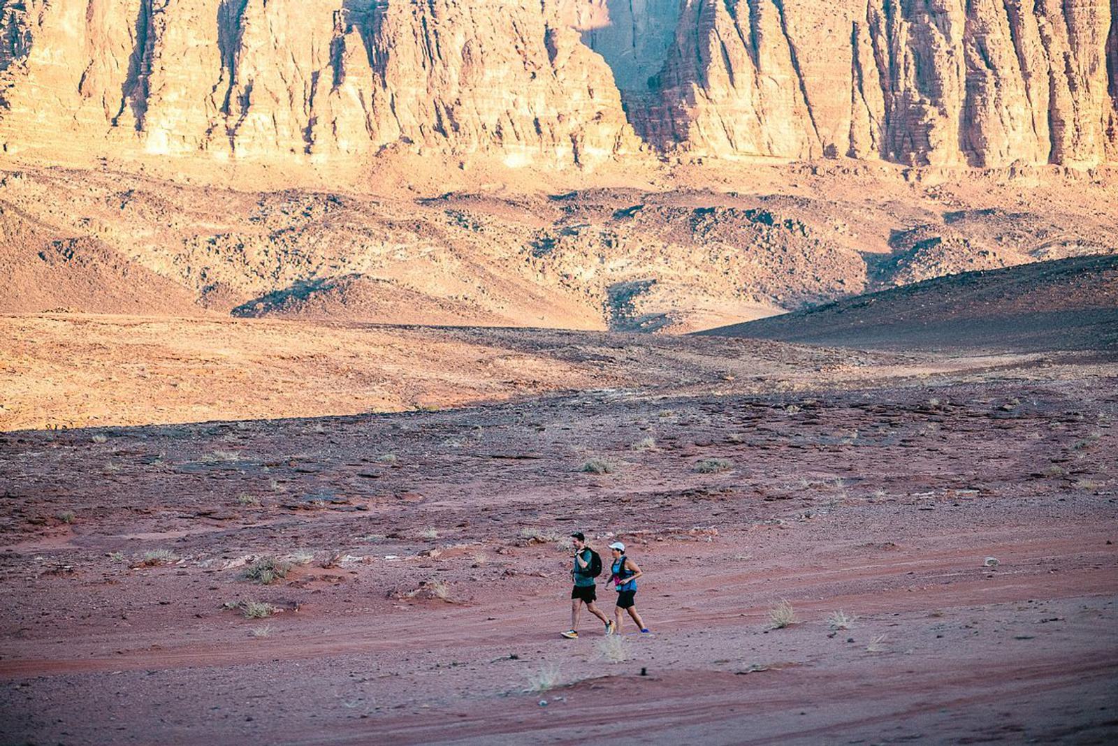 Two people are running through a vast, rocky desert landscape with towering cliffs in the background. The ground is arid, with patches of scattered shrubs, and the warm light accentuates the textures of the rugged terrain.