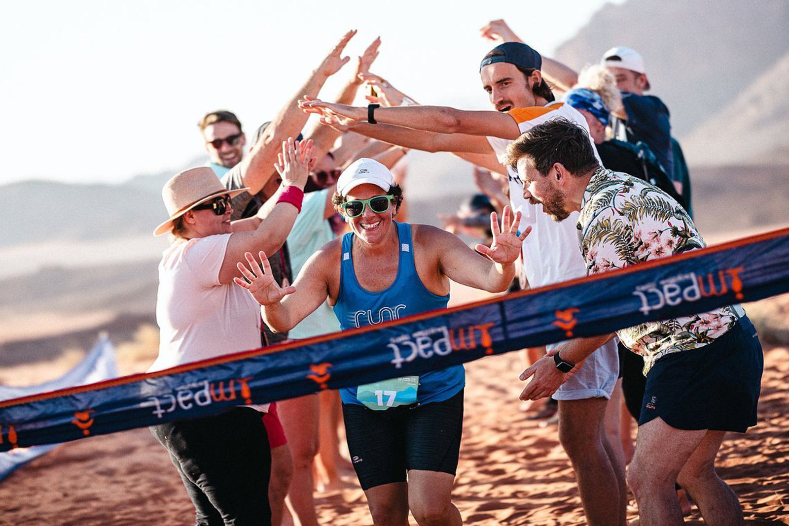 A smiling runner in a blue tank top and white cap high-fives cheering spectators as she crosses a finish line in a sandy, desert-like landscape. The spectators are enthusiastic, with arms raised, celebrating her achievement.
